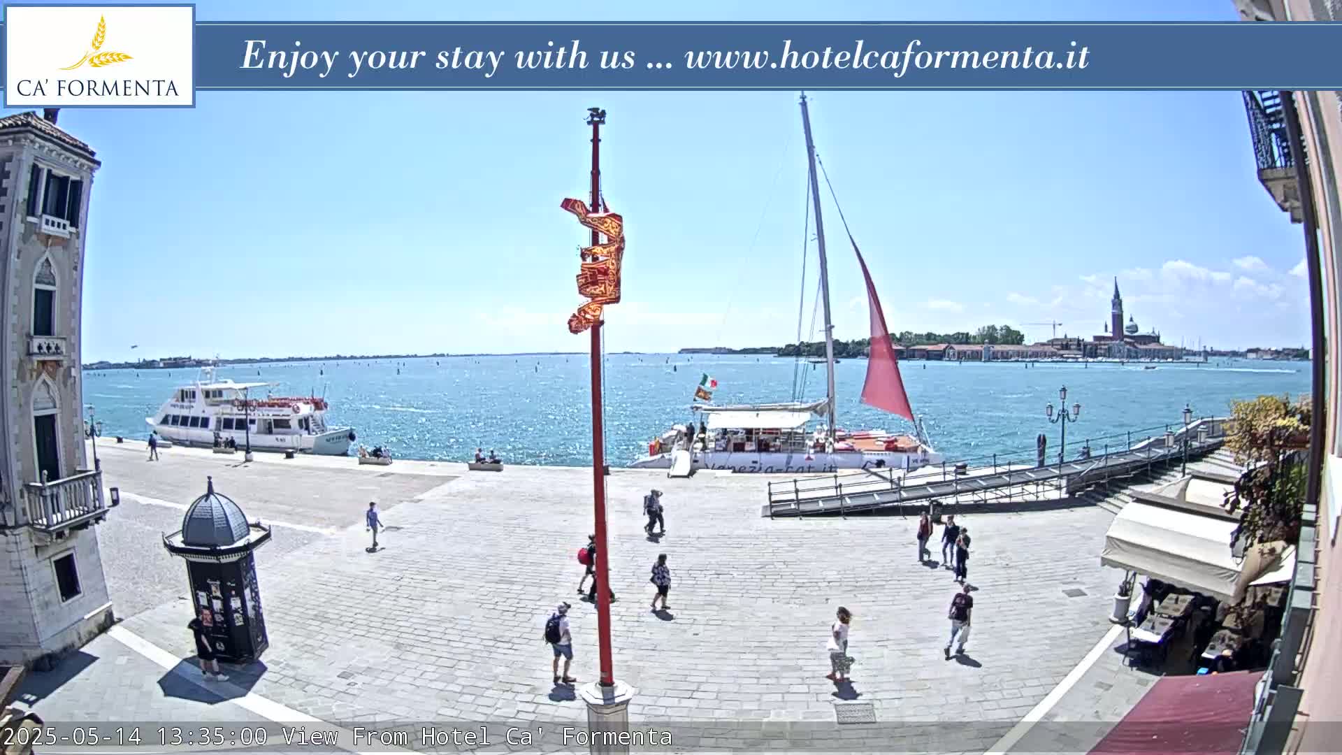 A sunny waterfront plaza with a few pedestrians, a docked tour boat, and a sailboat in the distance, overlooking a calm body of water and a distant island.