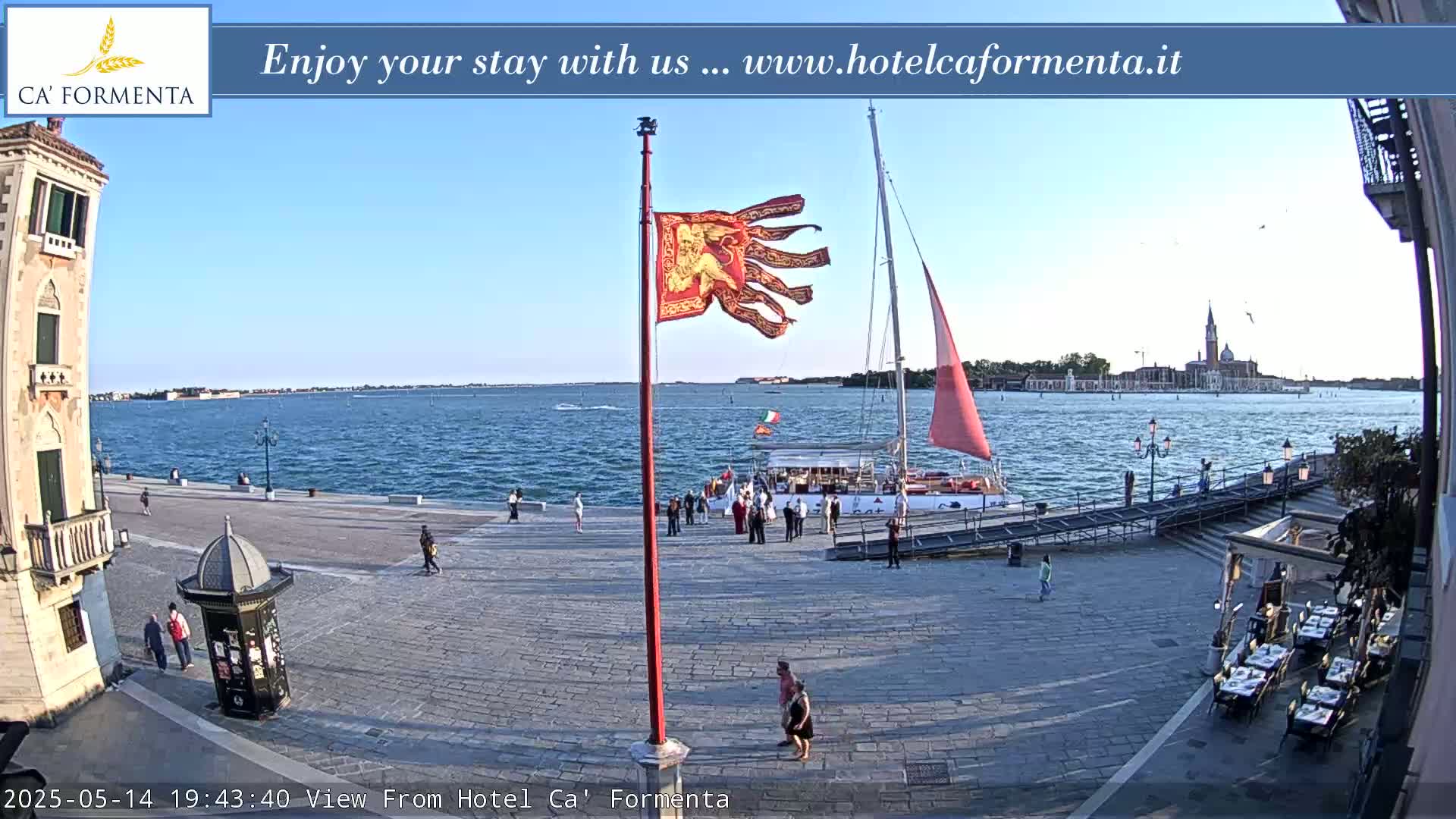 A waterfront plaza shows a sailboat at dock, a flagpole with a red banner, and several people, all under a clear, sunny sky.