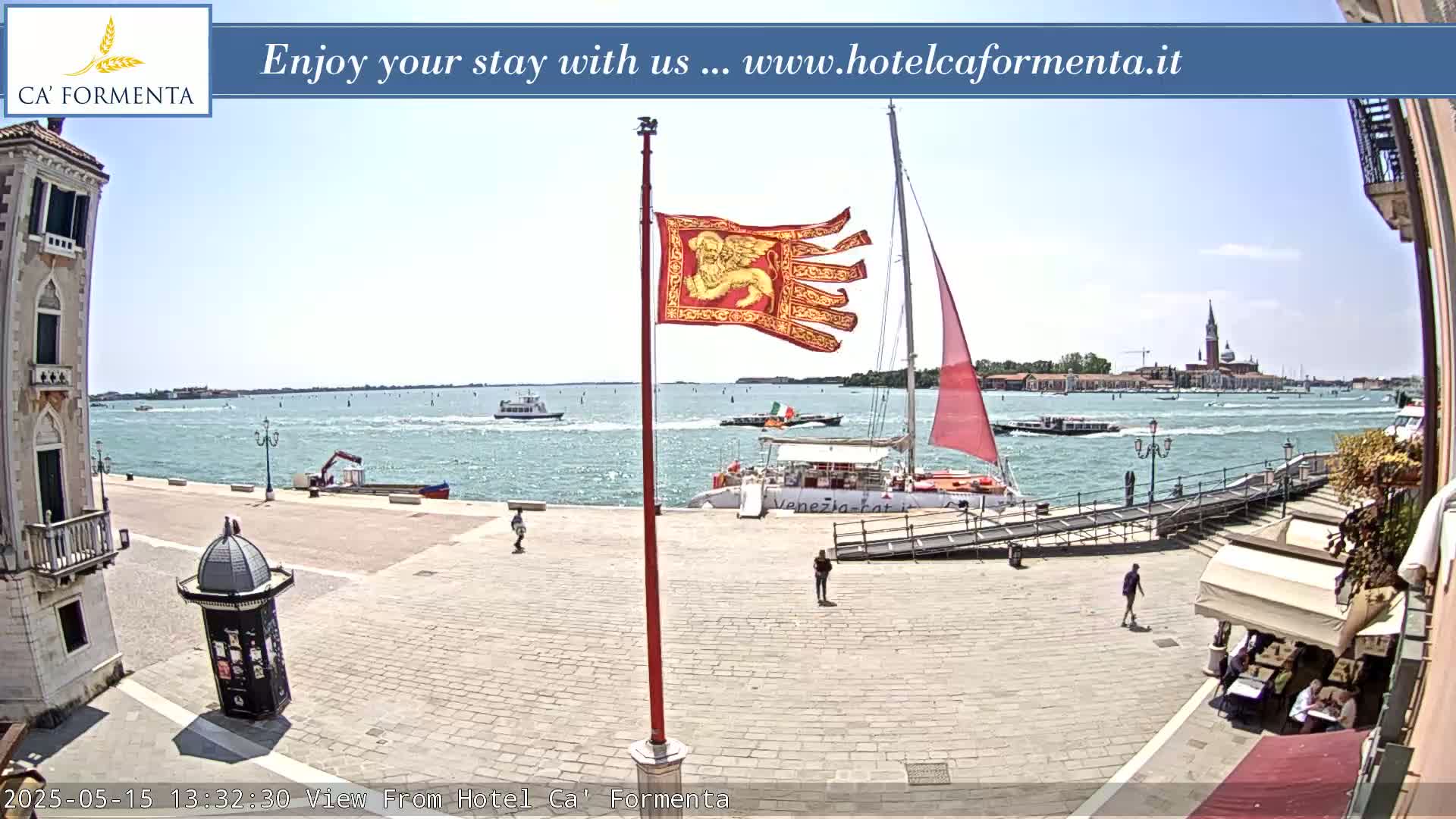 A sunny waterfront scene shows a paved plaza with a few people, a red sail boat docked at a gangway, a Venetian flag, and a calm body of water with other boats and a distant cityscape.