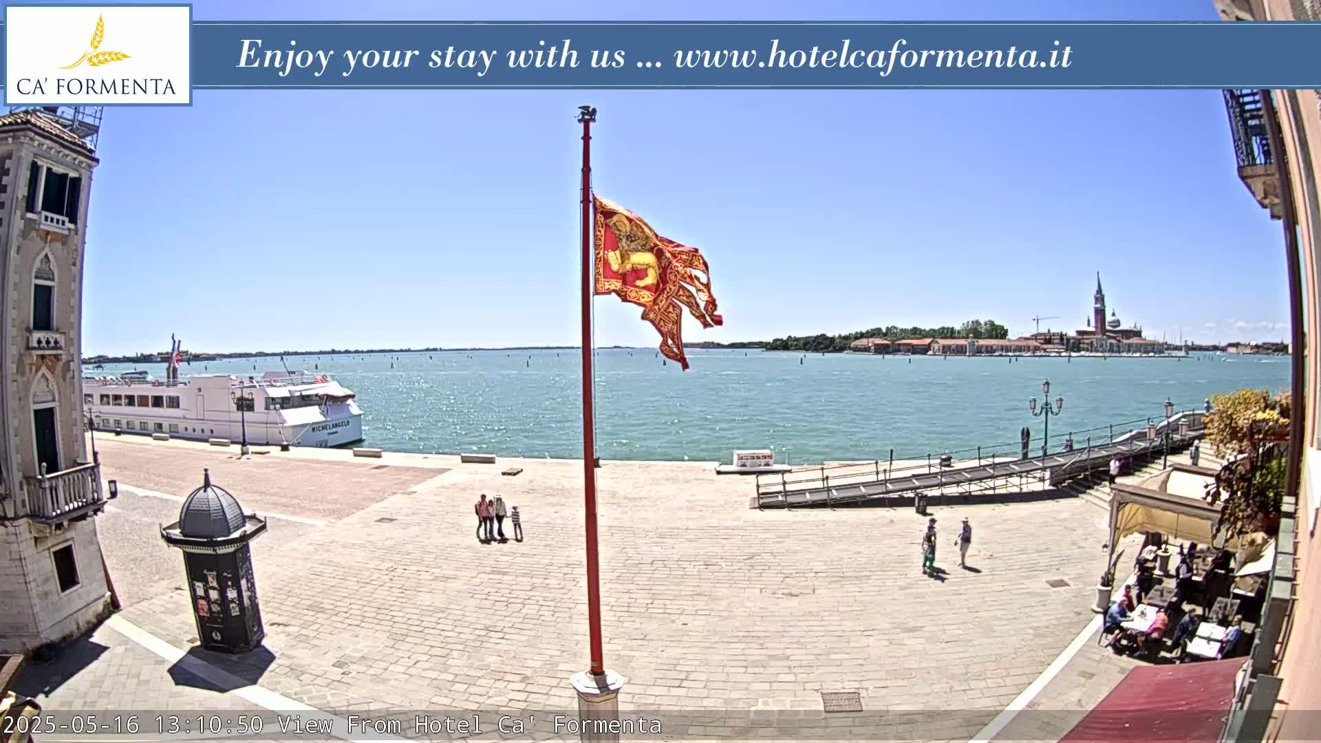 A sunny waterfront plaza shows a large cruise ship, a flagpole with a tattered flag, several people walking, and a distant island with buildings.