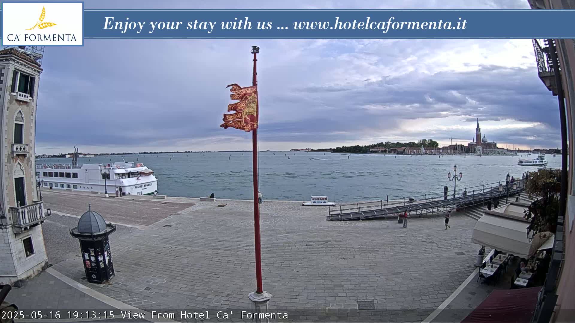 A waterfront cobblestone plaza overlooks a calm body of water with a boat and distant buildings under a cloudy sky.