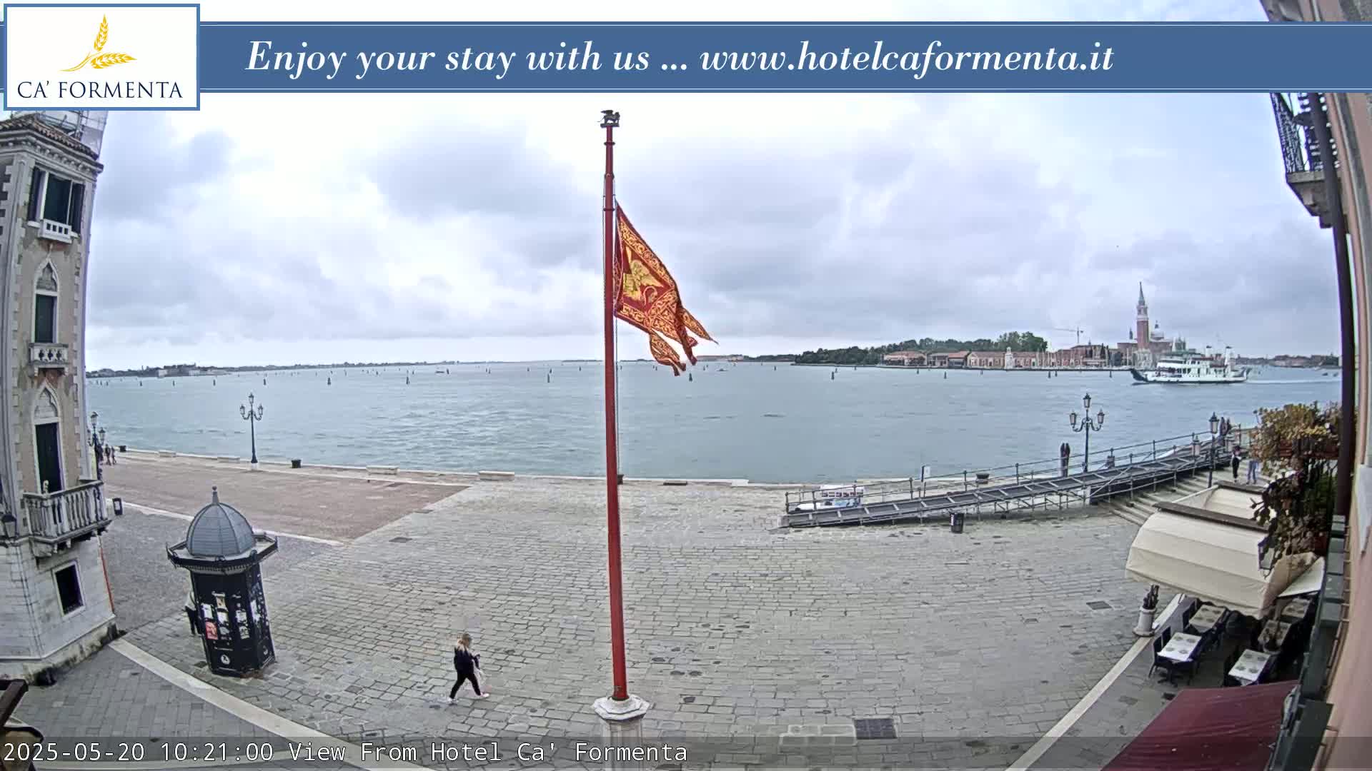 A mostly cloudy day overlooks a calm body of water with a boat and distant buildings, a red flagpole with a flag flies in the foreground near a paved area with a person walking.