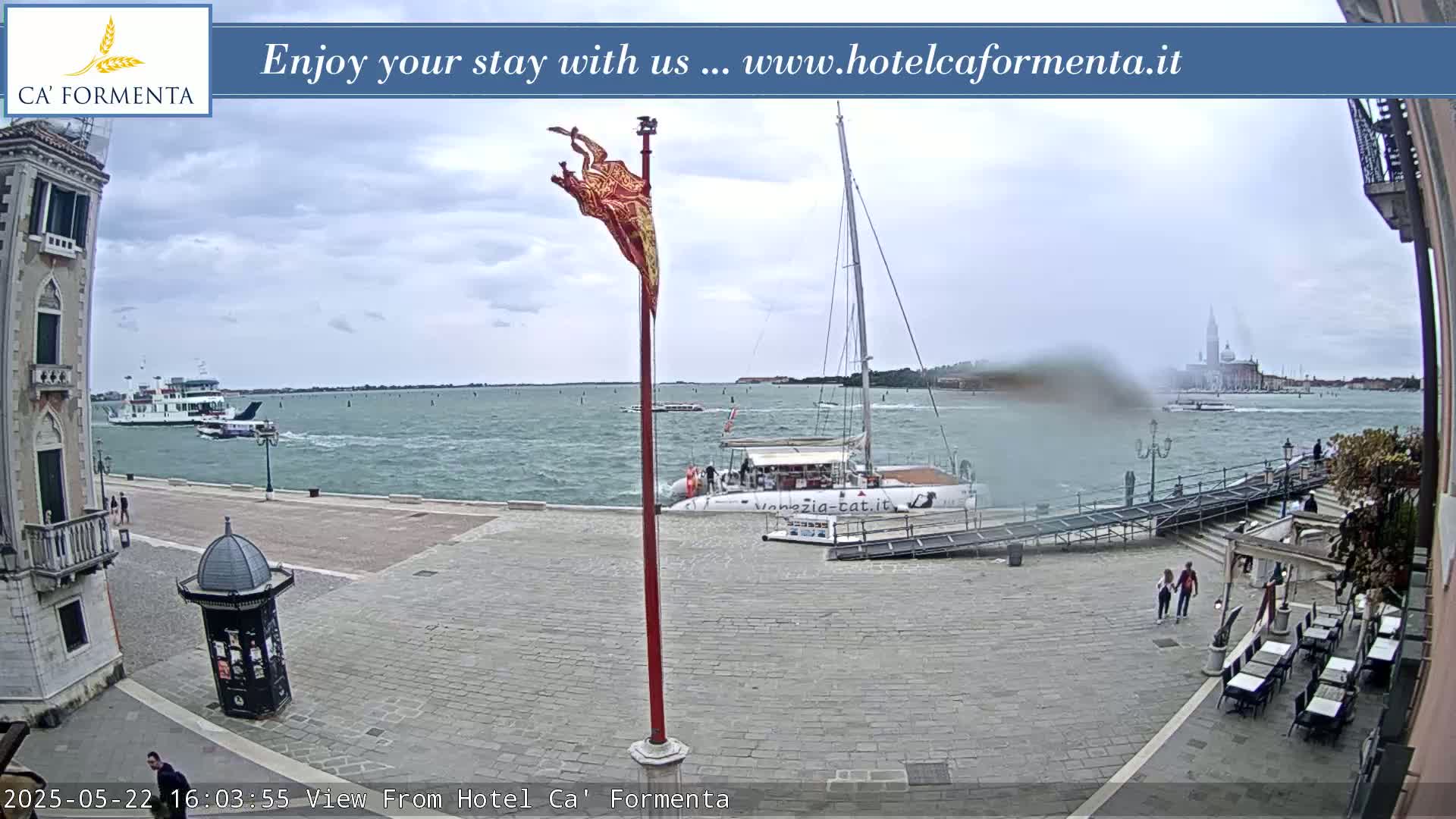 A waterfront plaza with a docked catamaran, a ferry in the distance, and overcast skies.