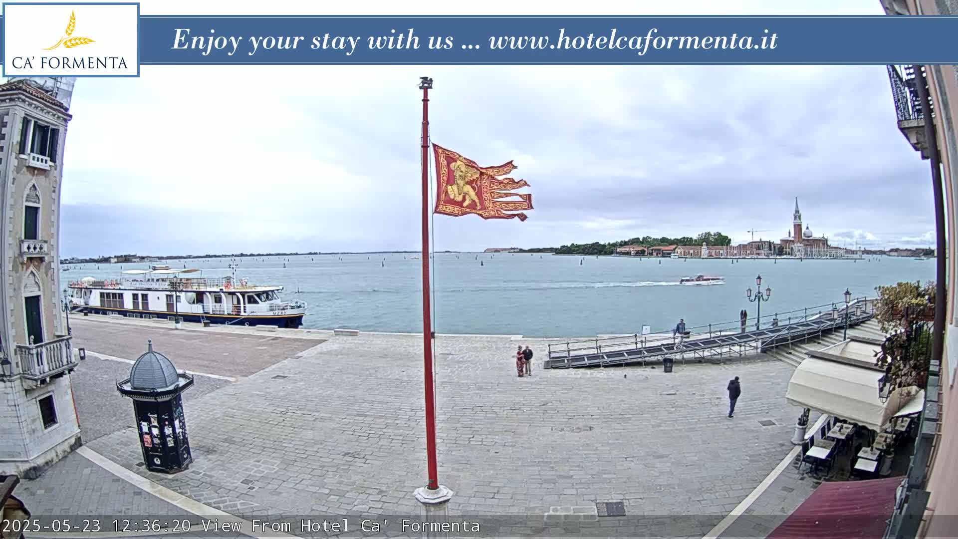 A waterfront paved plaza in overcast weather shows a boat docked nearby, with a distant view of buildings and a flagpole flying a tattered banner.