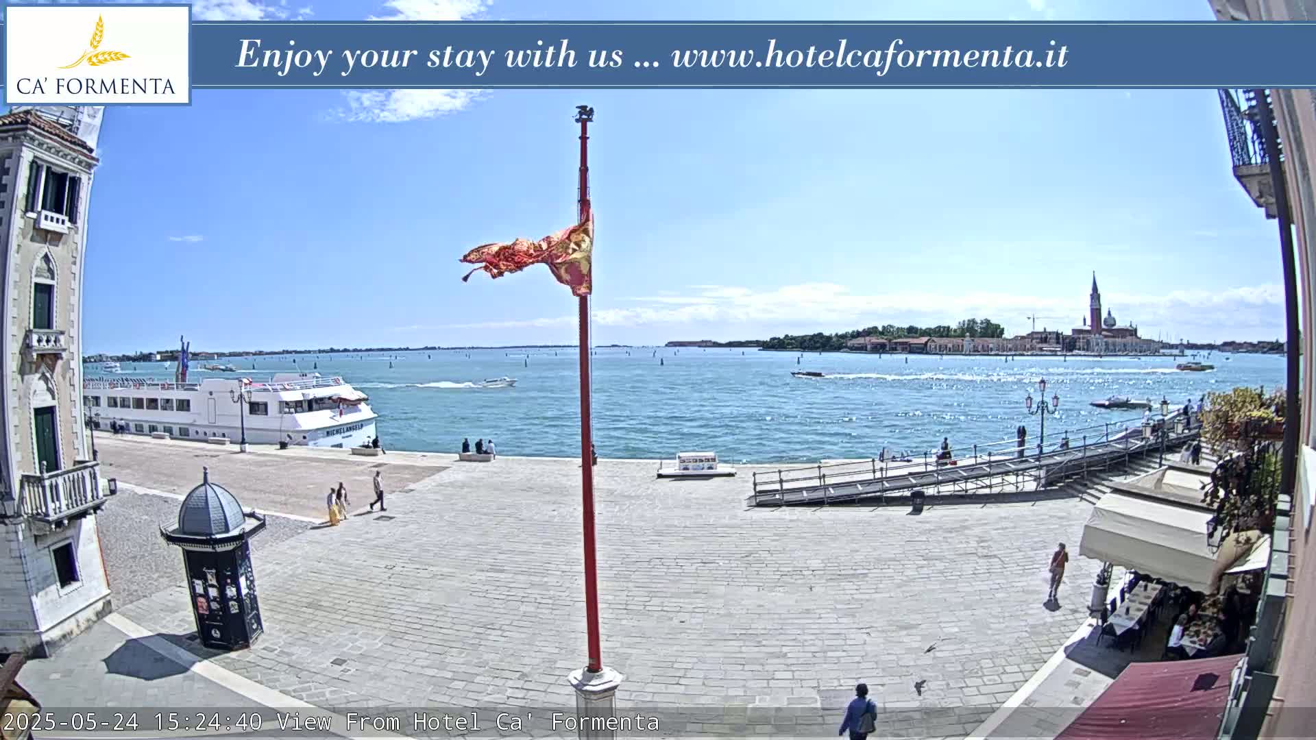 A sunny waterfront plaza overlooks a calm body of water with a large passenger ship and distant buildings under a clear sky.
