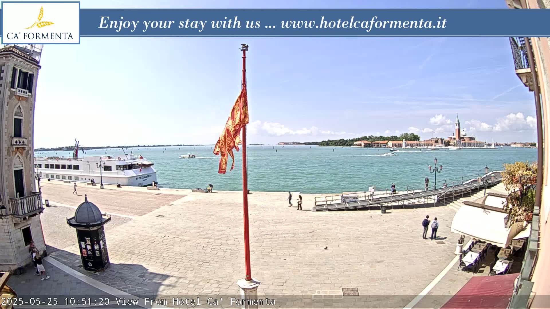 A waterfront plaza in sunny weather, showing a large cruise ship, a flagpole, and a distant view of a town across the water.