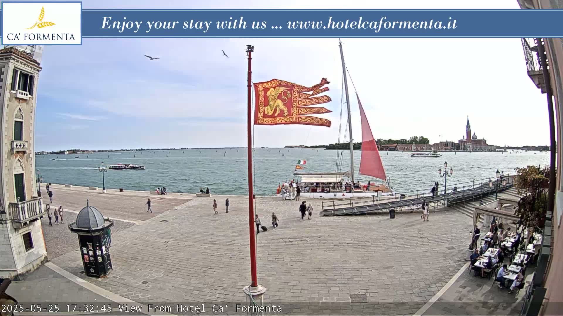 A sunny day in Venice shows a paved plaza with people walking, a boat docked near a flagpole with a red banner, and a view of the water with boats and buildings in the distance.