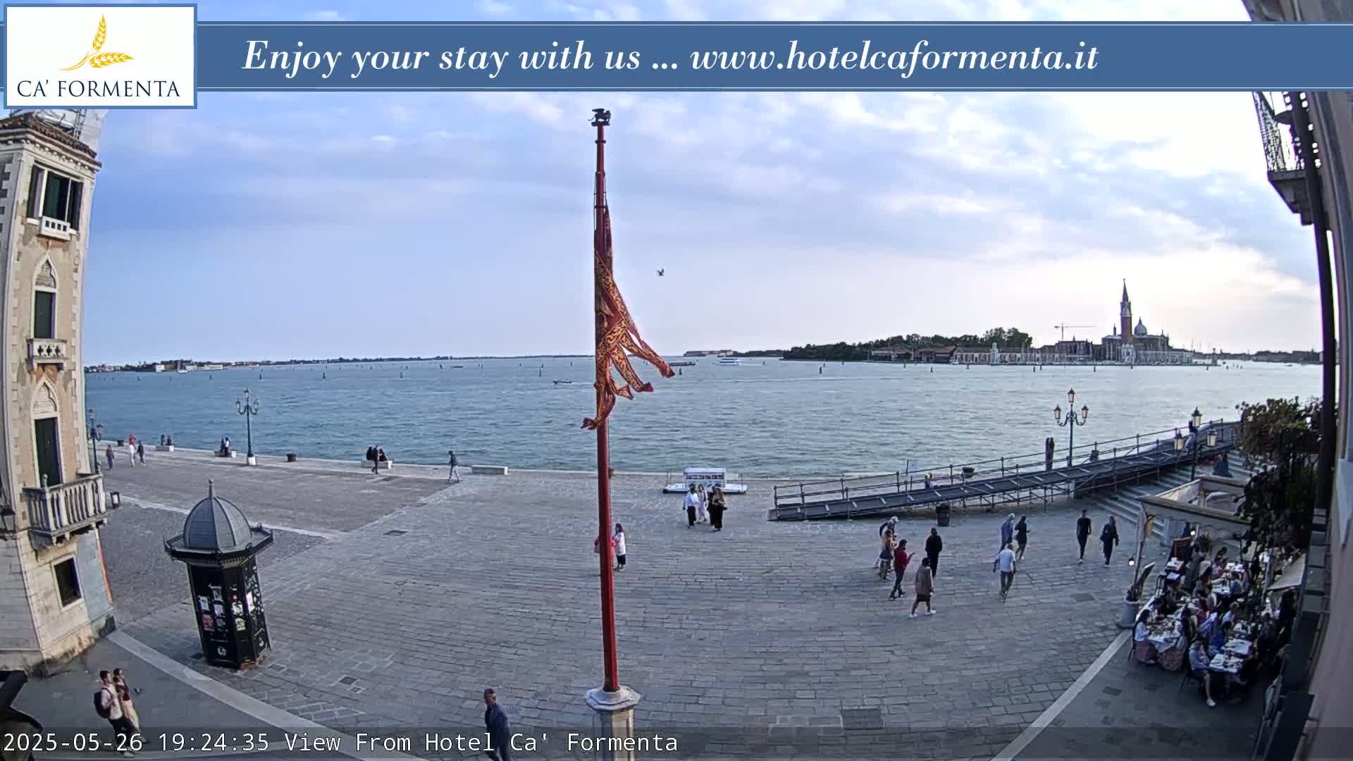 A partly cloudy outdoor scene shows a paved plaza overlooking a calm body of water with distant buildings and a flagpole with a hanging banner.