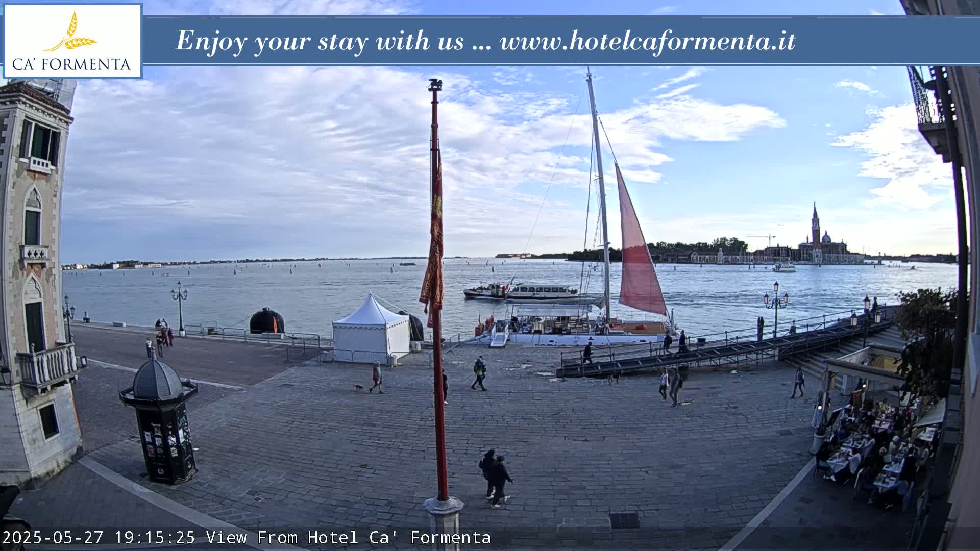 A waterfront paved area with a sailboat docked, a few people walking around, and a body of water showing a distant cityscape under a partly cloudy sky.