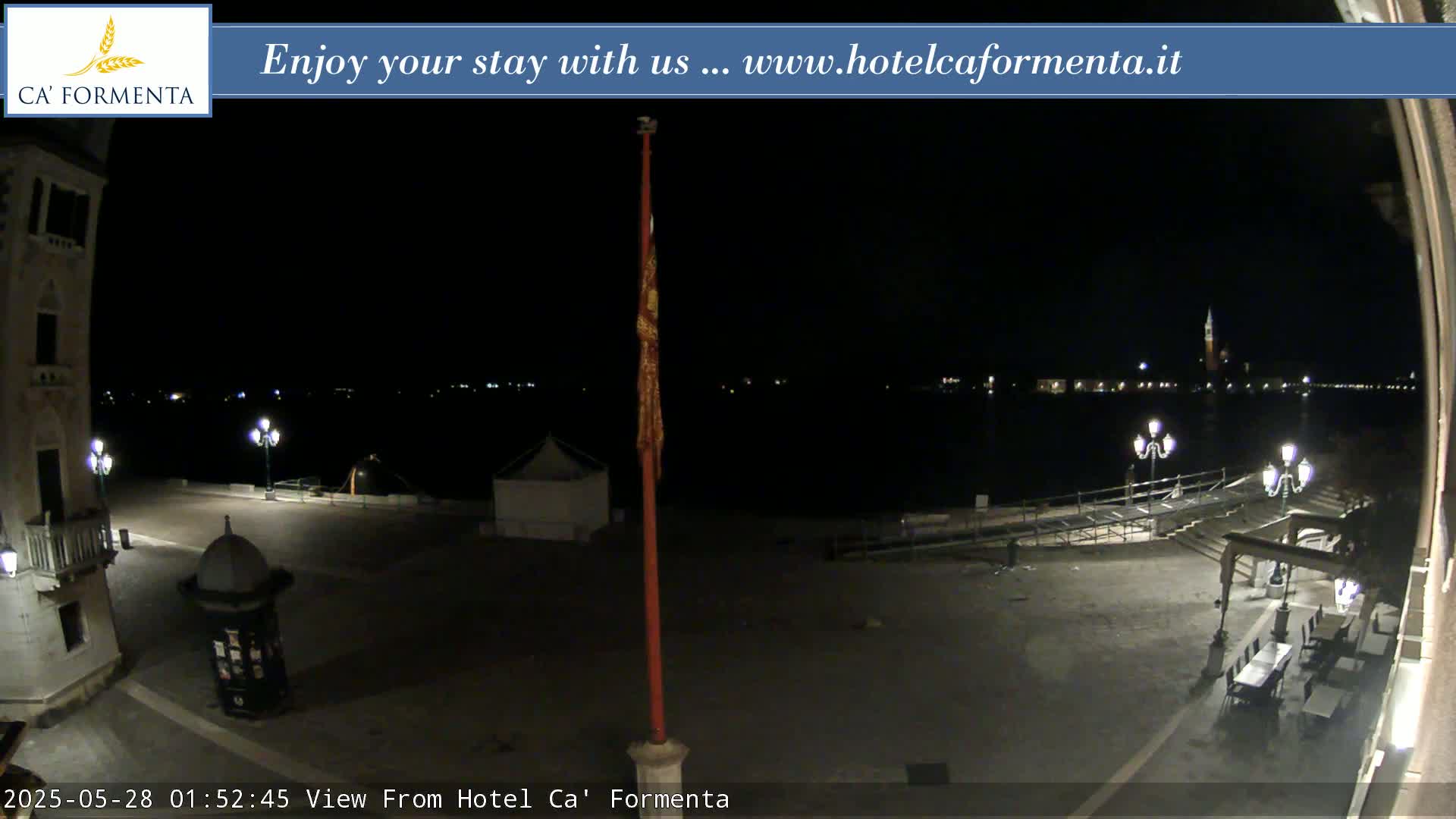 A nighttime view of a plaza overlooking a dark body of water with distant lights, under a clear sky.