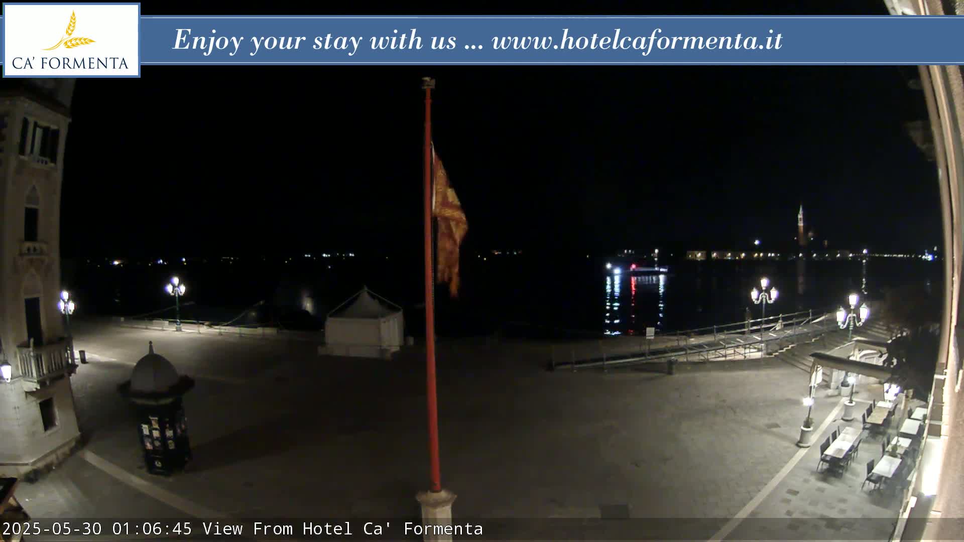 A nighttime view of a waterfront plaza with a flagpole, outdoor seating, and a body of water with distant lights.
