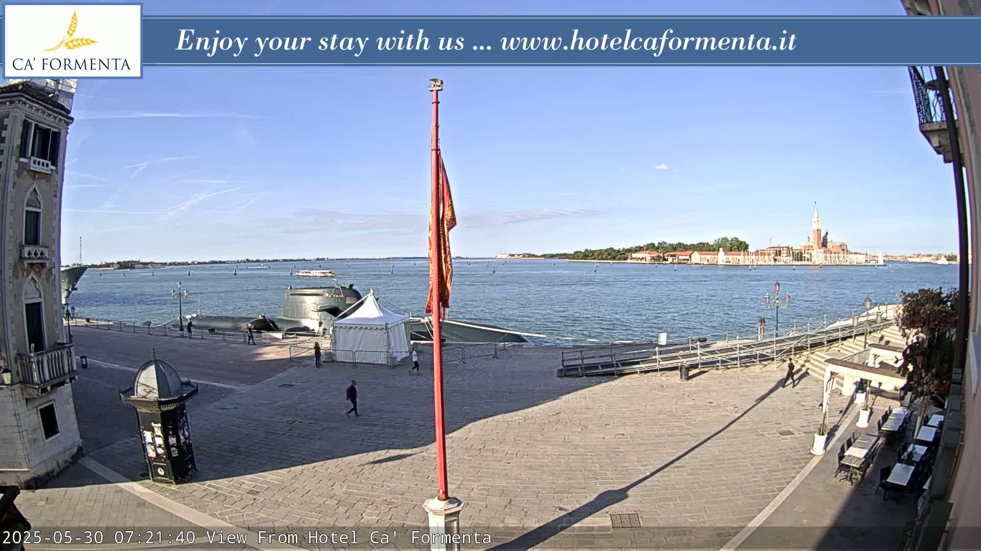 A sunny day overlooks a calm body of water with a distant cityscape, a partially visible submarine, and a paved plaza with a few people and outdoor seating.