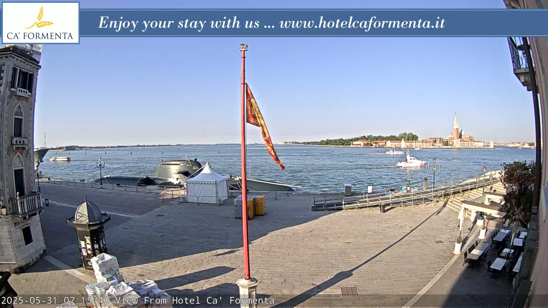 A sunny outdoor scene shows a paved plaza overlooking a calm body of water with boats and a distant cityscape under a clear blue sky.