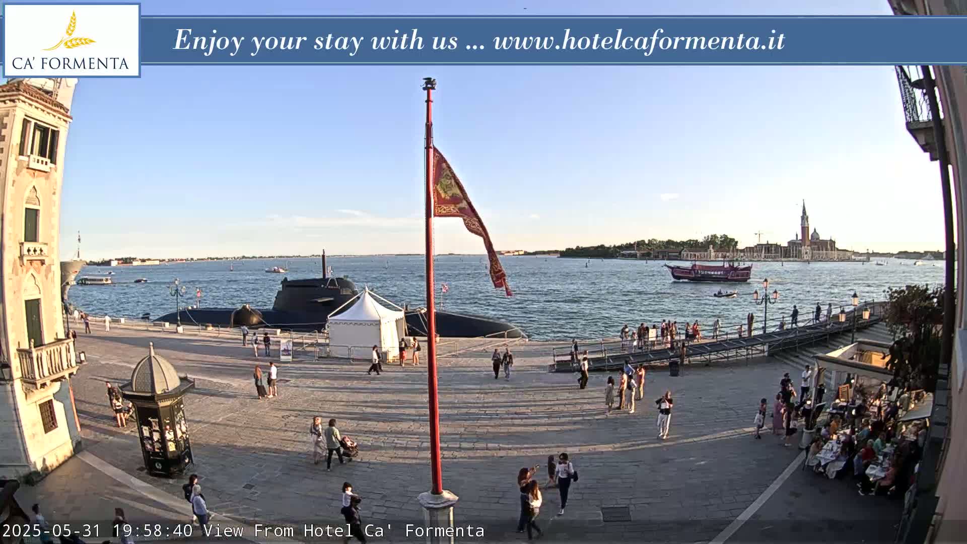 A submarine is docked near a waterfront plaza filled with people, under a clear, sunny sky, with a large red boat visible in the water beyond.