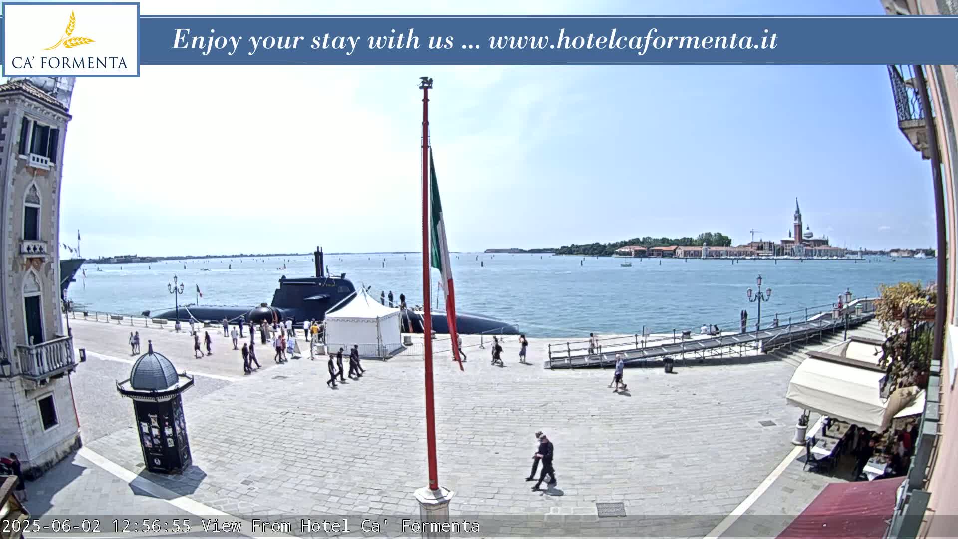 A submarine is docked near a waterfront plaza on a sunny day, with people walking around and a distant view of an island.