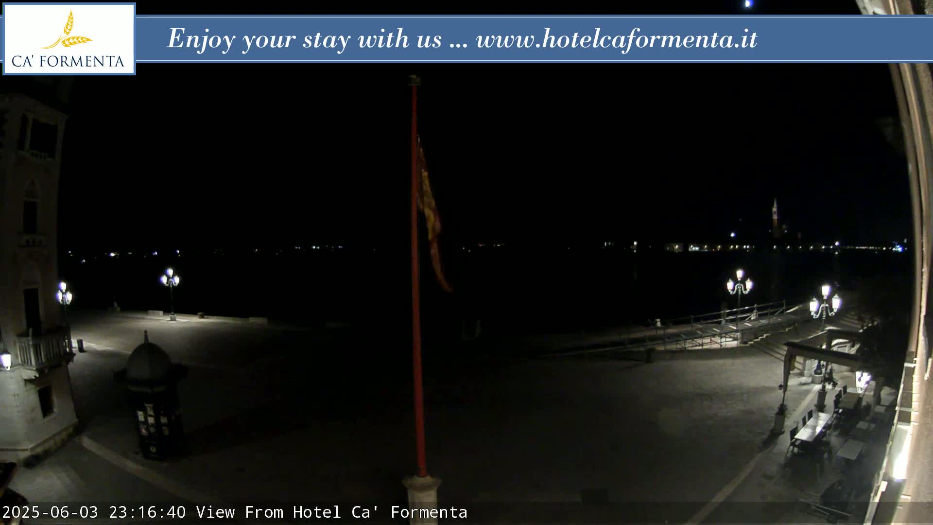 A nighttime view of a dark, mostly empty plaza with a flagpole and distant city lights across water.