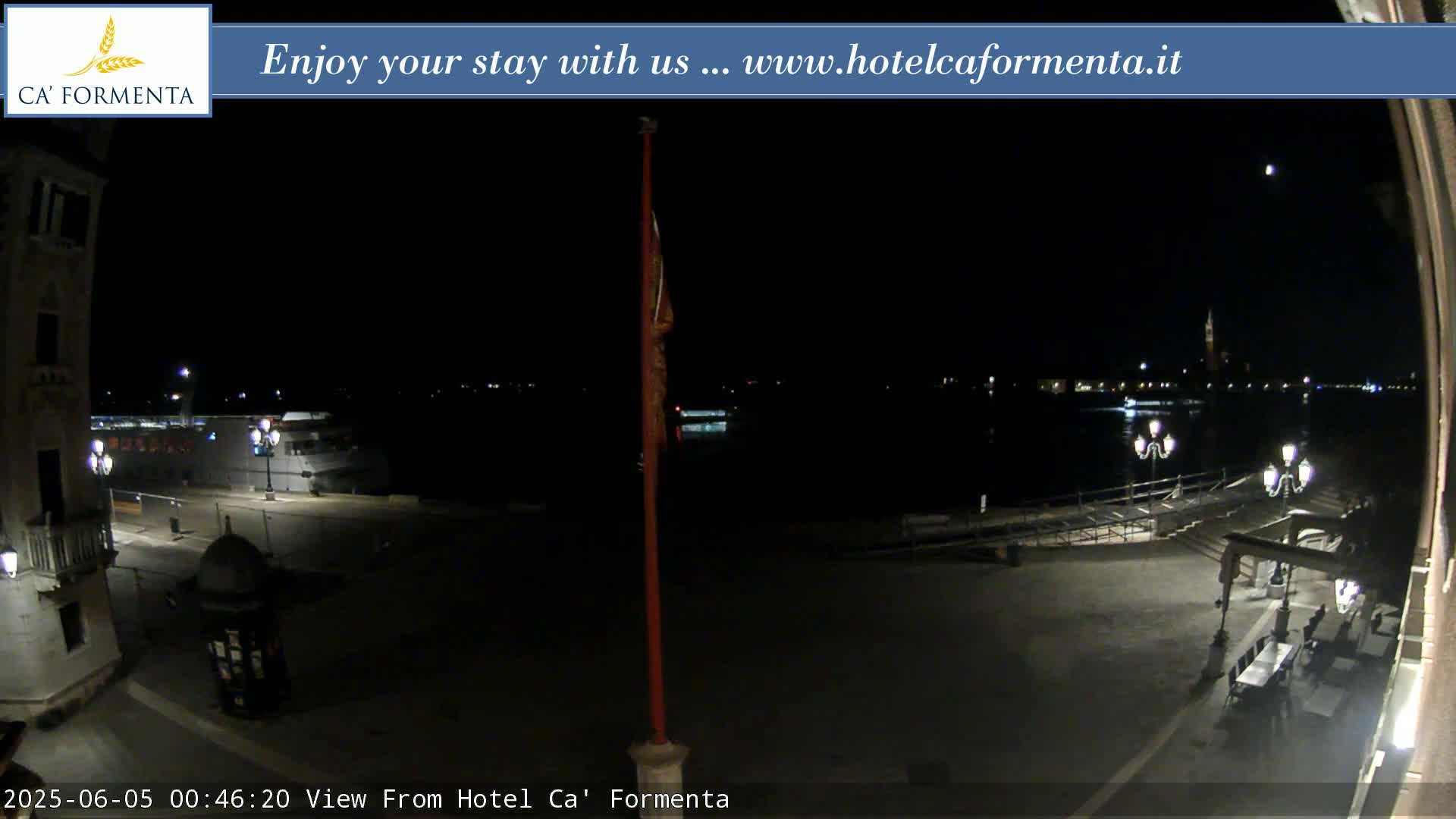 A nighttime view of a waterfront plaza shows a dark body of water, a few lights across the water, and some outdoor seating near a building.