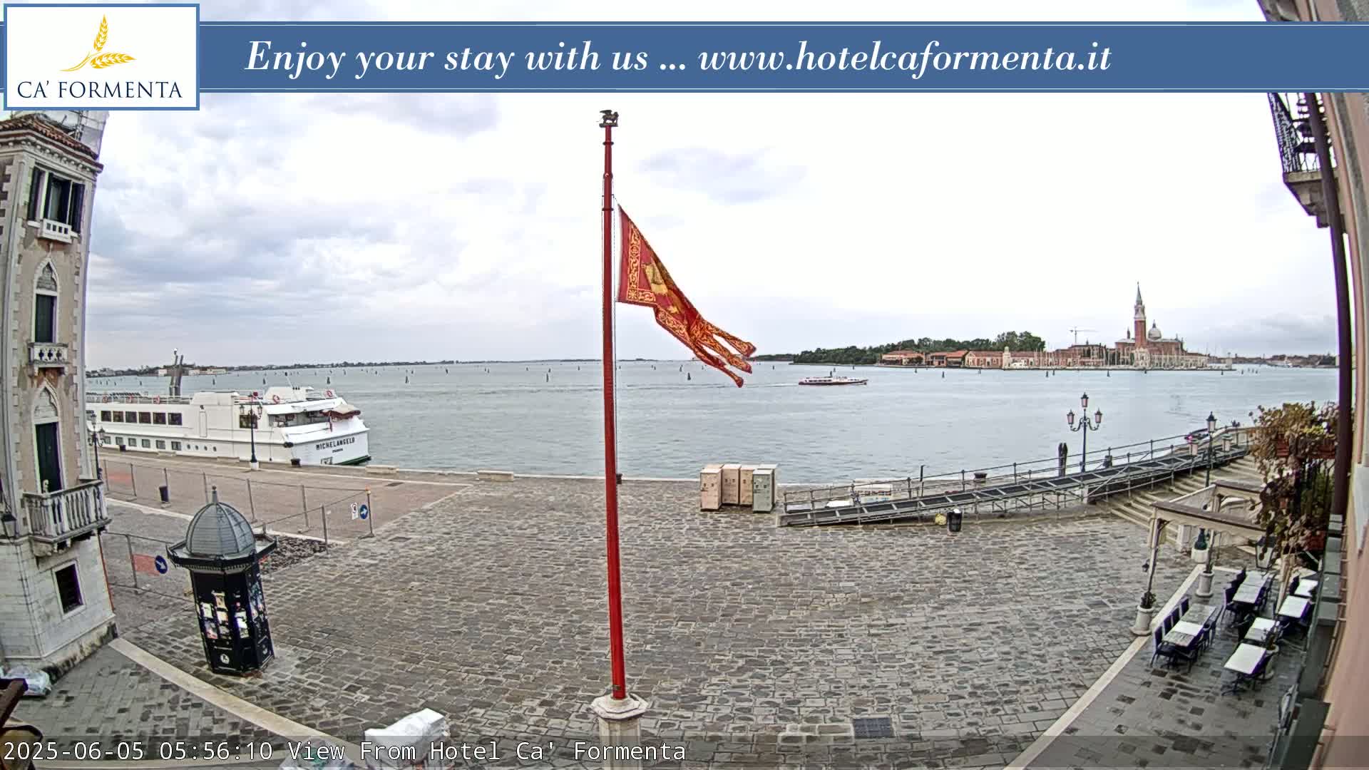 A paved waterfront square overlooks a calm body of water with a large cruise ship and distant buildings under an overcast sky, a red flag flying on a tall pole.