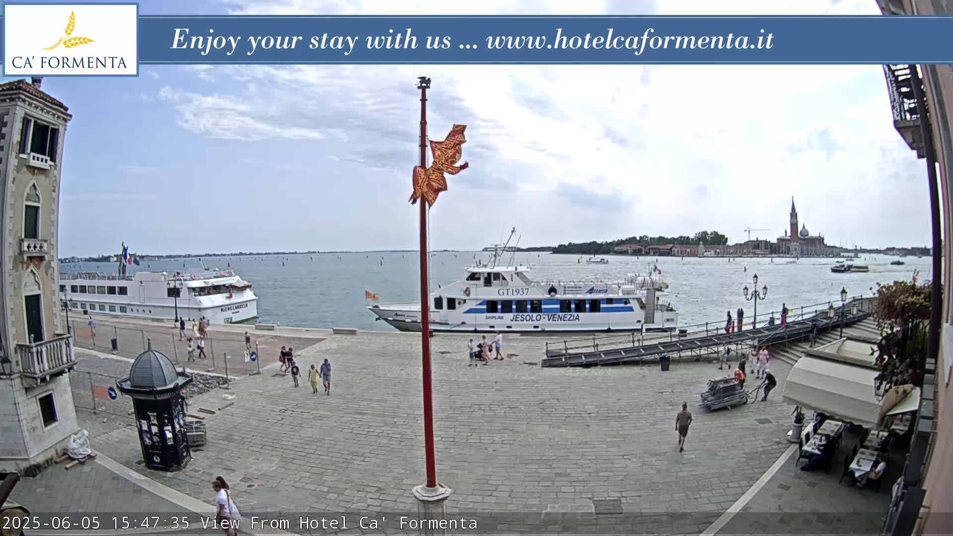 A partly cloudy day shows a waterfront paved plaza with several people, two docked boats, and a distant view of a city across calm water.