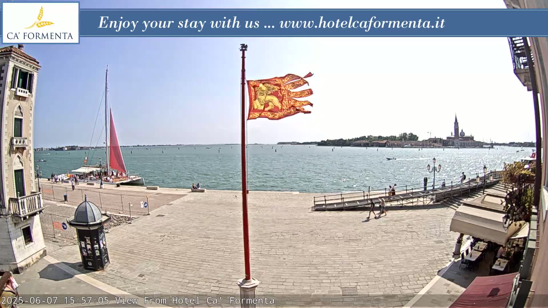 A sunny waterfront scene shows a paved plaza with a red flagpole, a red sailboat in the water, and a distant island with buildings.