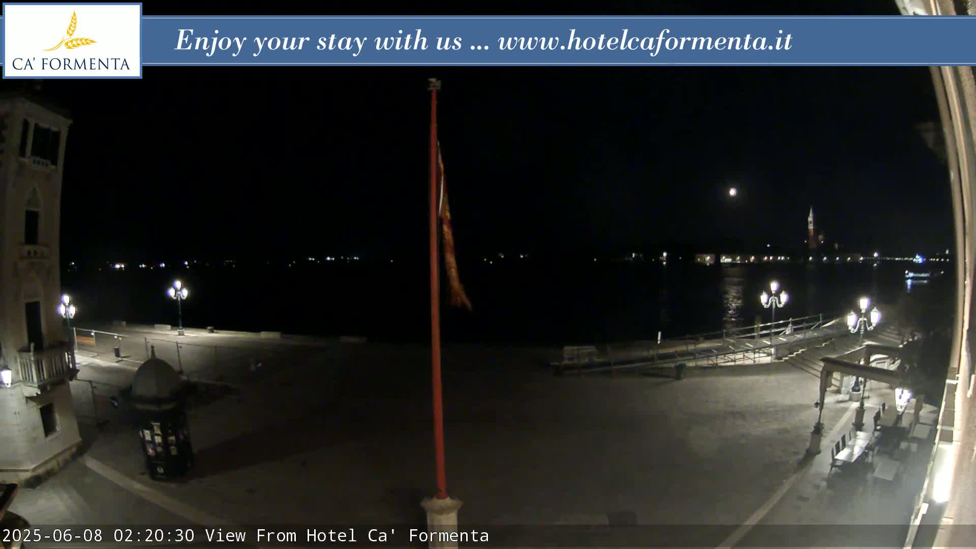 A nighttime view of a waterfront plaza shows a calm body of water with lights across the far shore under a slightly moonlit sky.