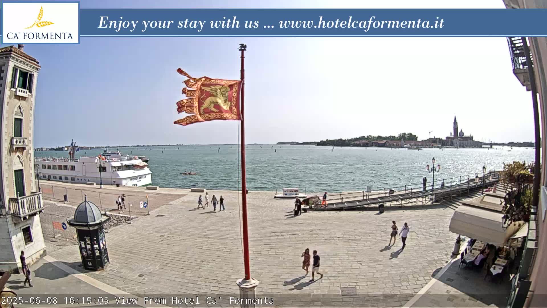 A sunny waterfront scene shows a paved plaza with pedestrians, a docked cruise ship, and a distant island with buildings under a clear sky.