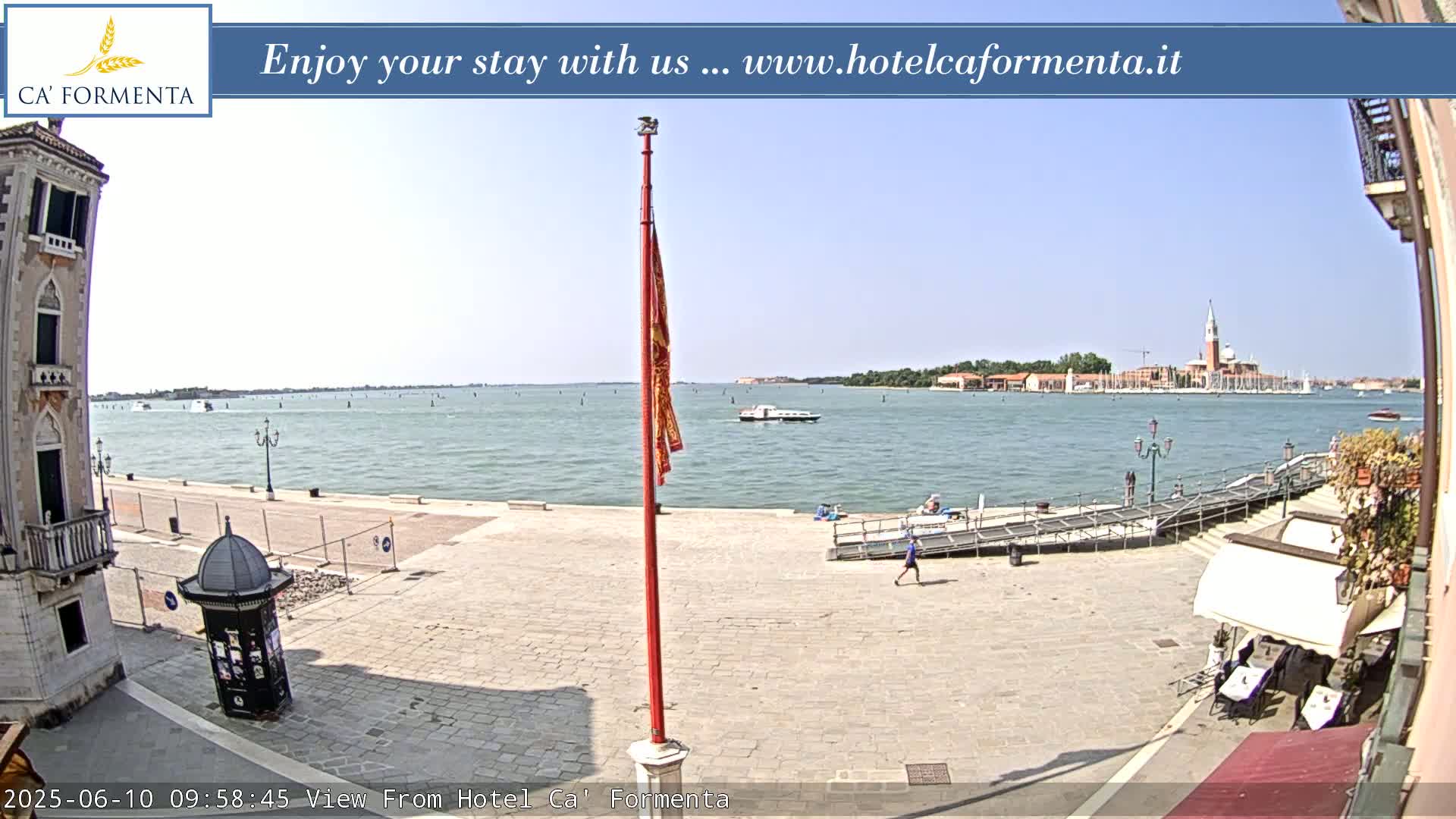 A sunny Venetian cityscape shows a paved plaza overlooking a calm body of water with a distant island and a few boats.