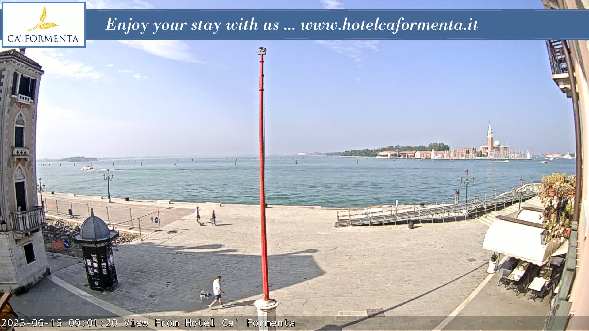 A sunny waterfront view shows a paved plaza with a few pedestrians, a tall red pole, and a distant island with buildings visible across calm water.