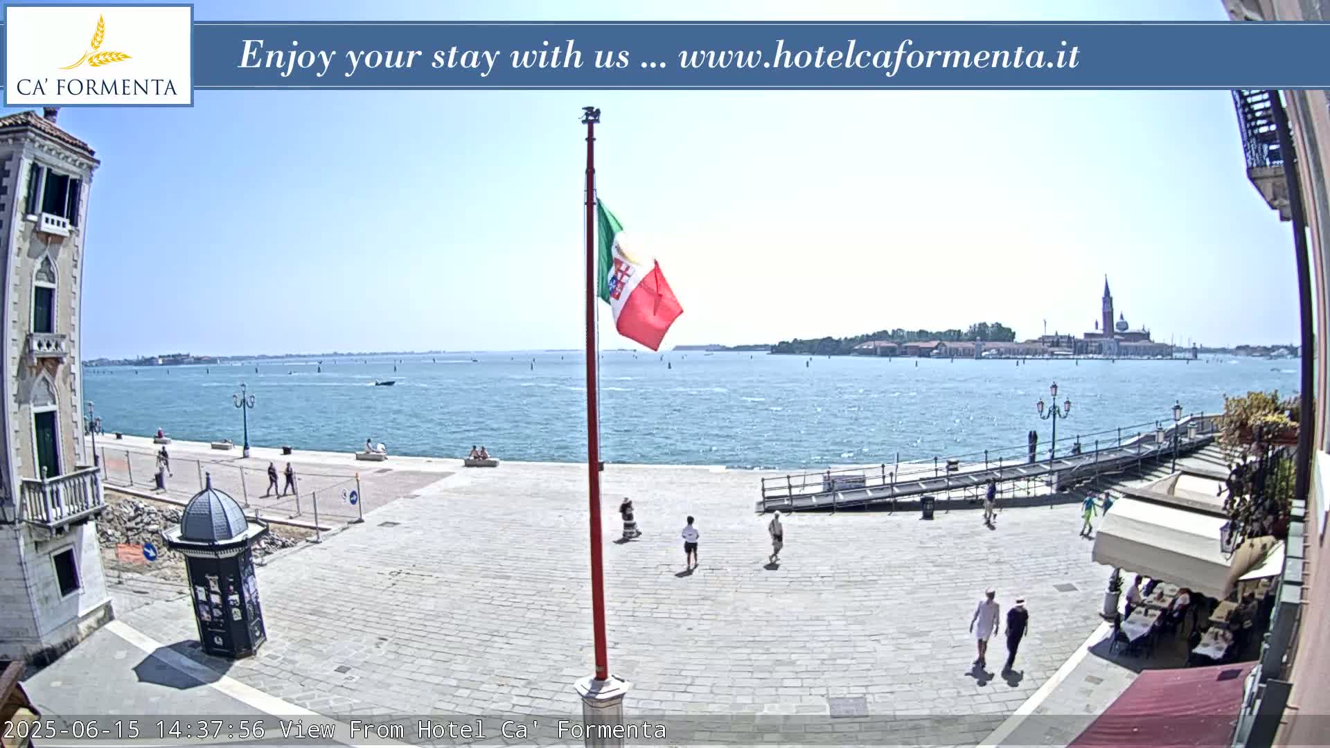 A sunny outdoor scene shows a paved plaza overlooking a calm body of water with distant buildings and a flagpole displaying an Italian flag.