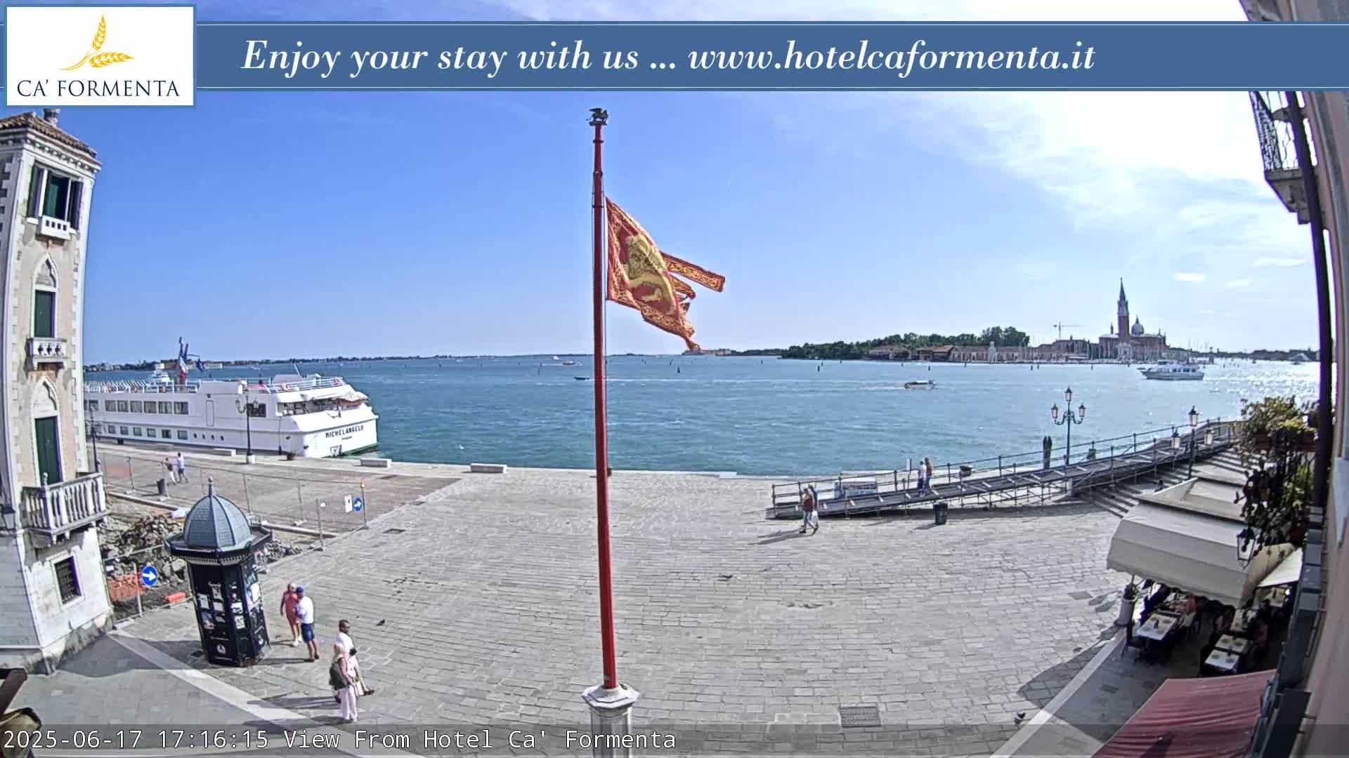 A sunny waterfront scene shows a large cruise ship docked near a paved plaza with several people walking, a flagpole with a tattered flag, and a distant view of buildings across the water.