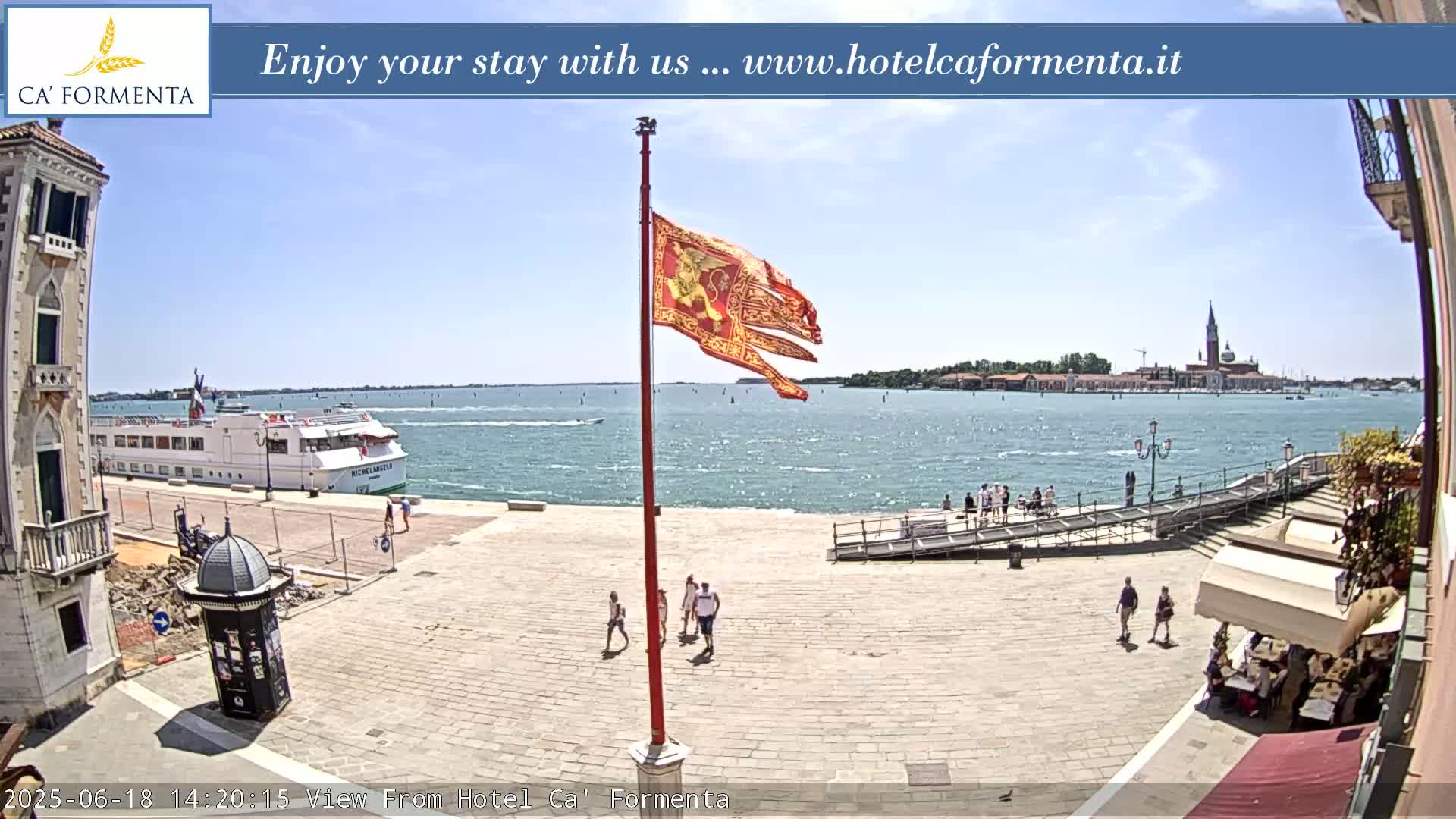A sunny waterfront plaza scene shows a flagpole with a tattered red flag, a cruise ship in the water, and a few pedestrians walking near a restaurant with an outdoor seating area.