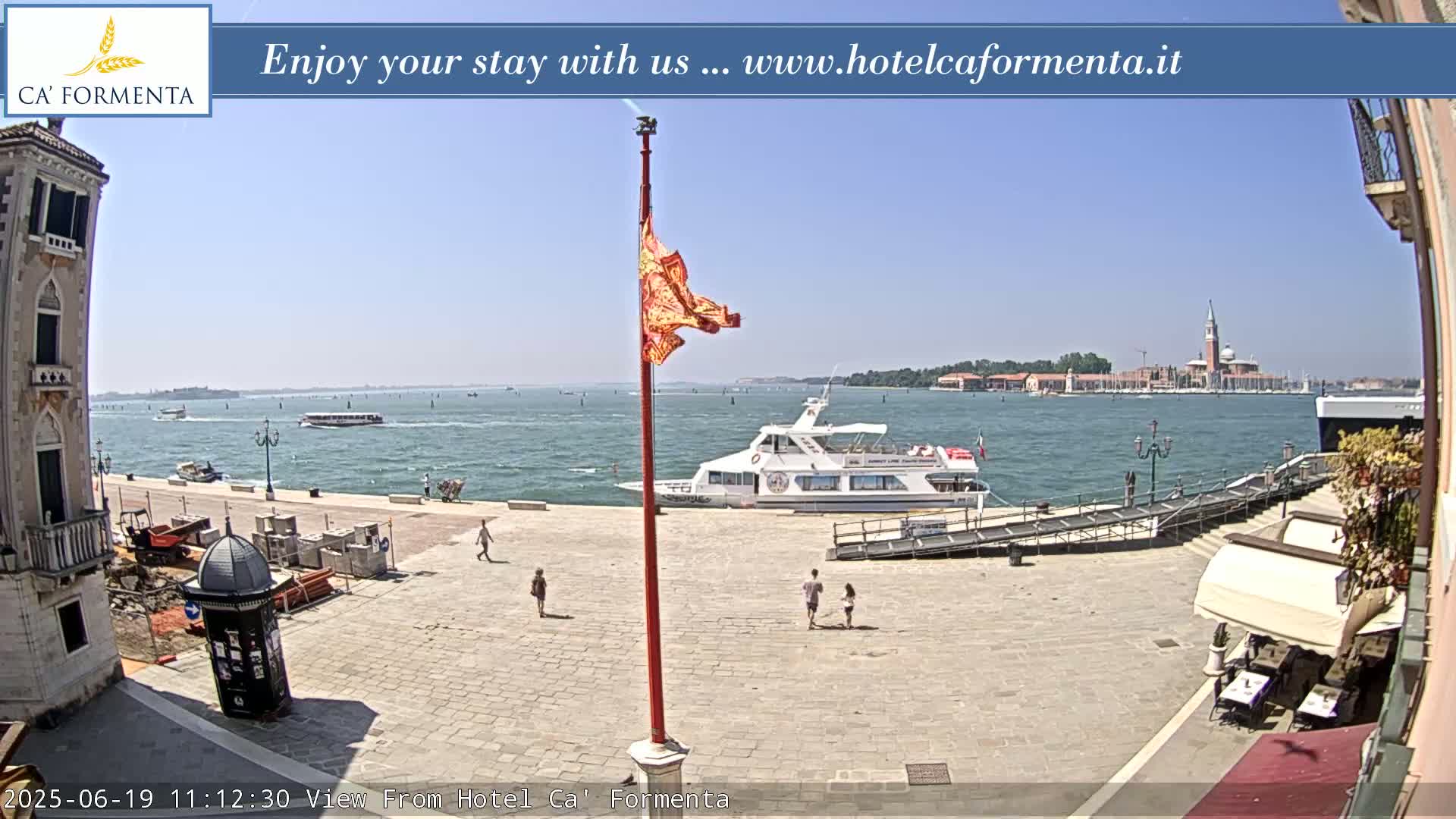 A sunny day in Venice shows a paved plaza with a few people walking, construction materials, and a waterfront with a large passenger boat and distant islands.