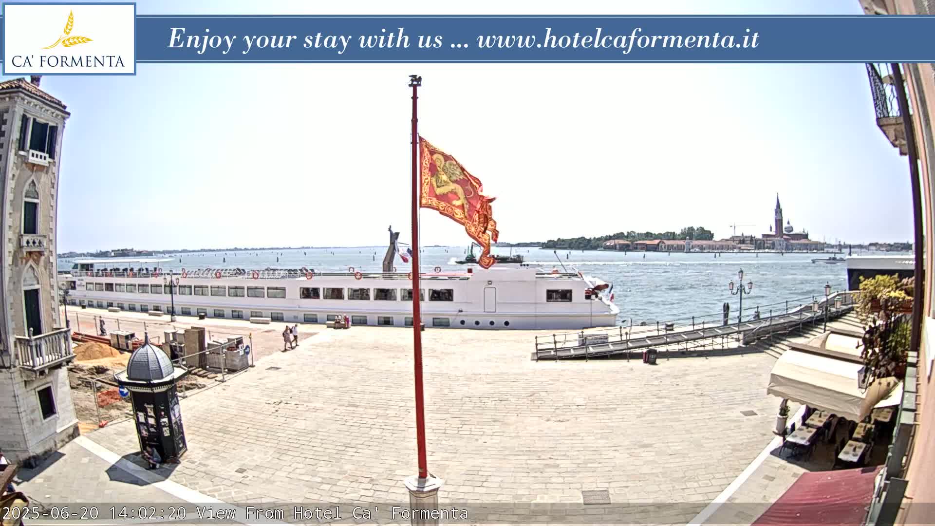 A large passenger ship is docked near a paved plaza, with a flagpole displaying a red flag and a distant view of a city across calm water under a clear sky.