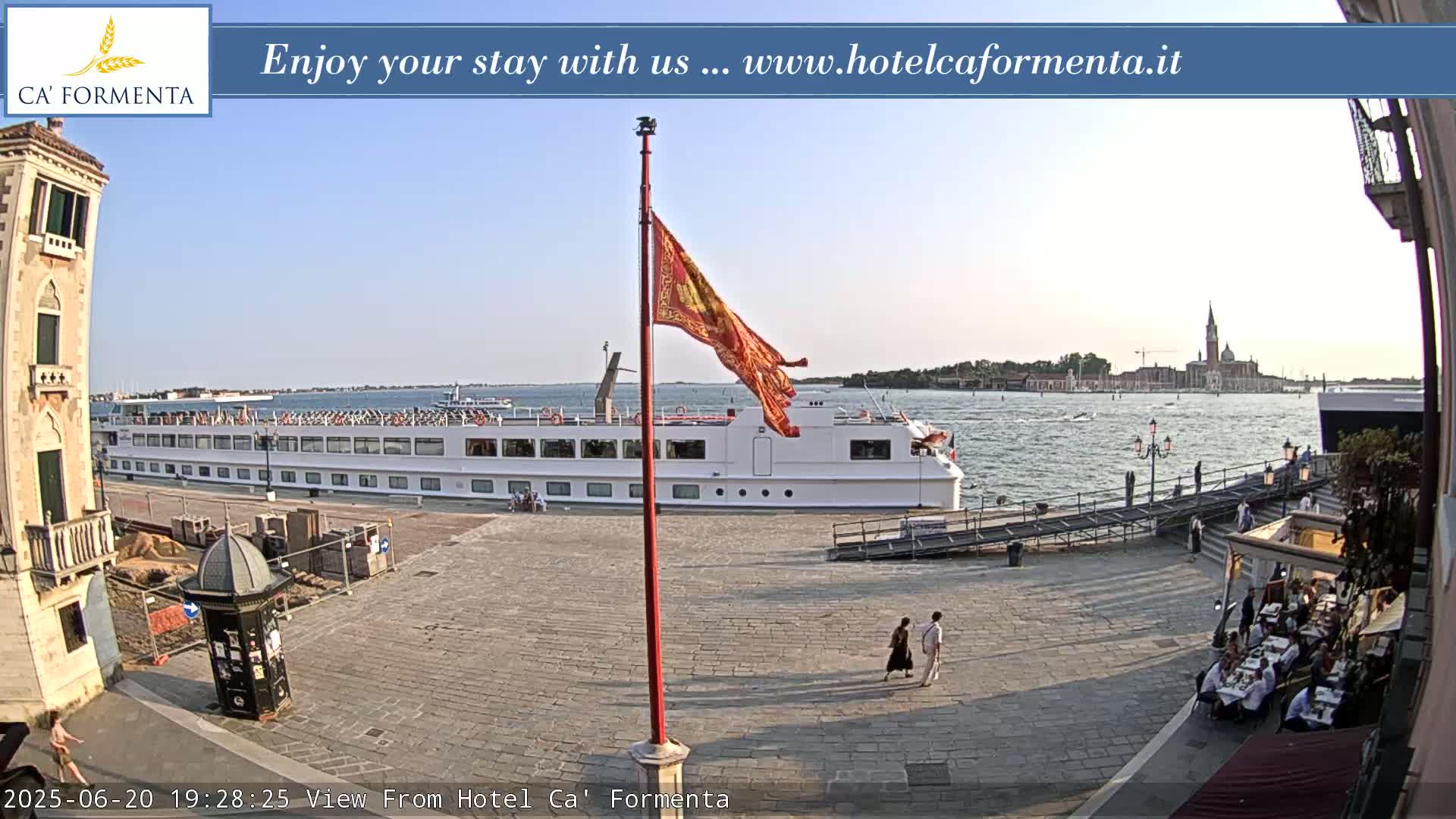 A large white passenger ship is docked near a paved area with people walking and dining outdoors, under a clear, sunny sky.