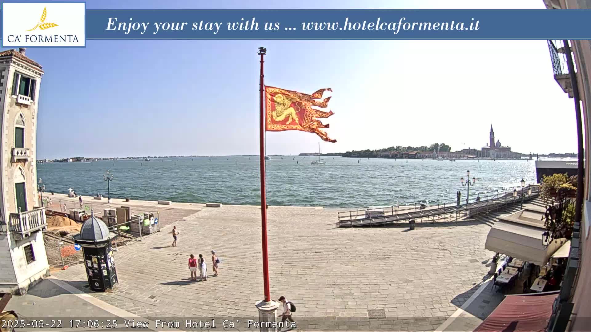 A sunny outdoor scene shows a paved plaza overlooking a calm body of water with a distant cityscape and a tattered red flag on a pole in the foreground.