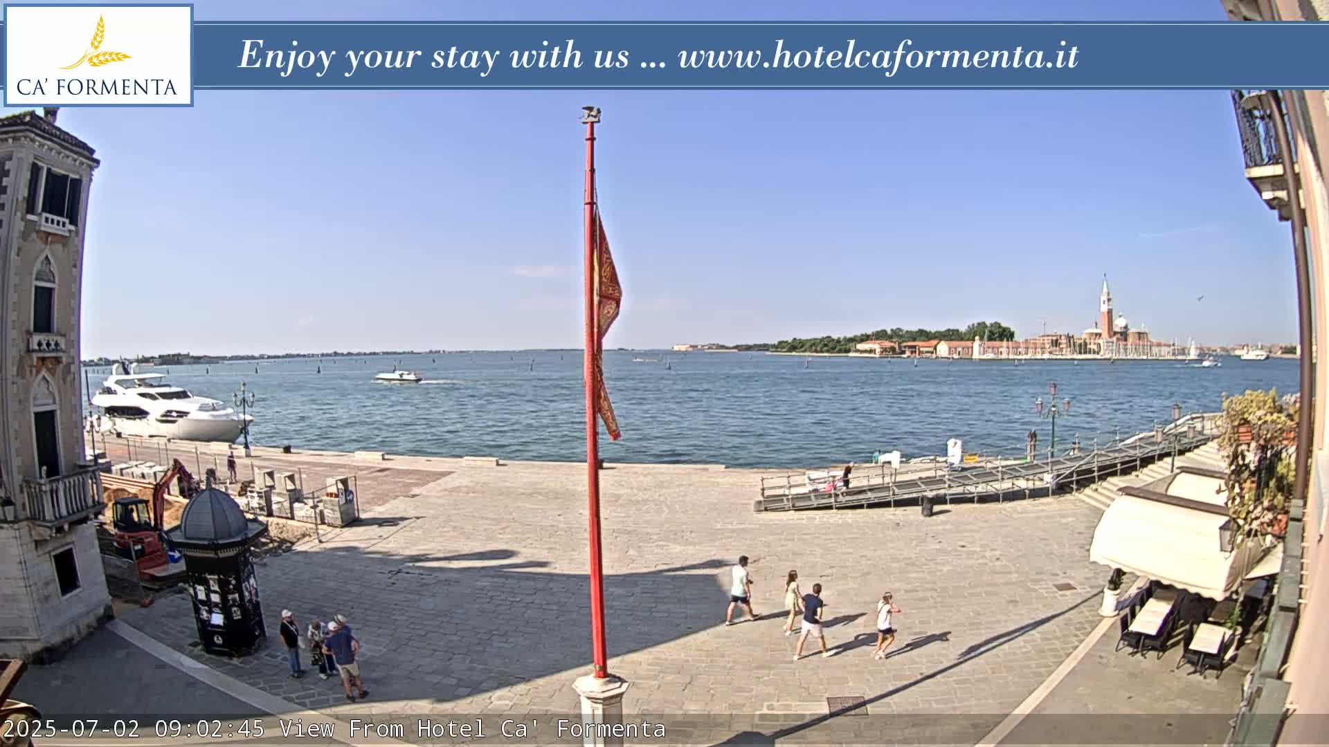 A sunny waterfront plaza shows several people walking, a yacht in the water, and a distant island with buildings.