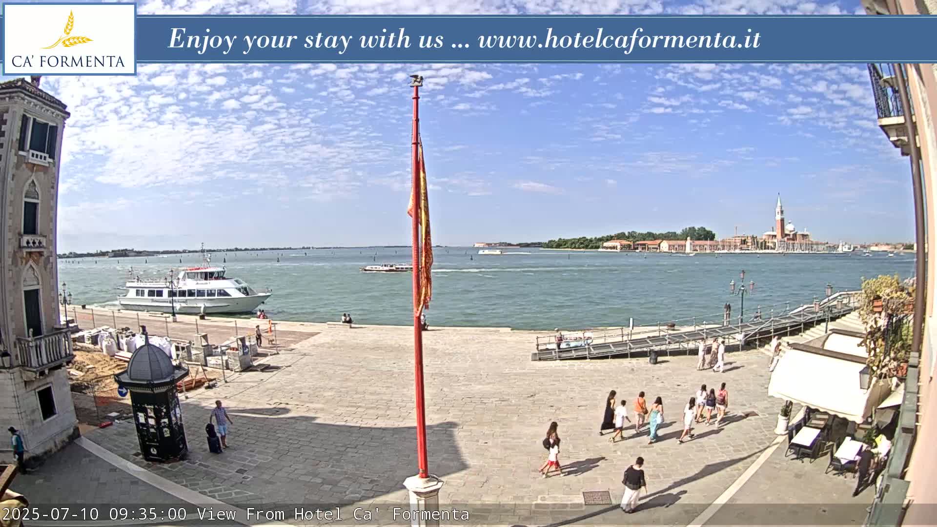 A sunny day shows a waterfront plaza with people walking, a boat docked in the water, and a distant island with buildings.