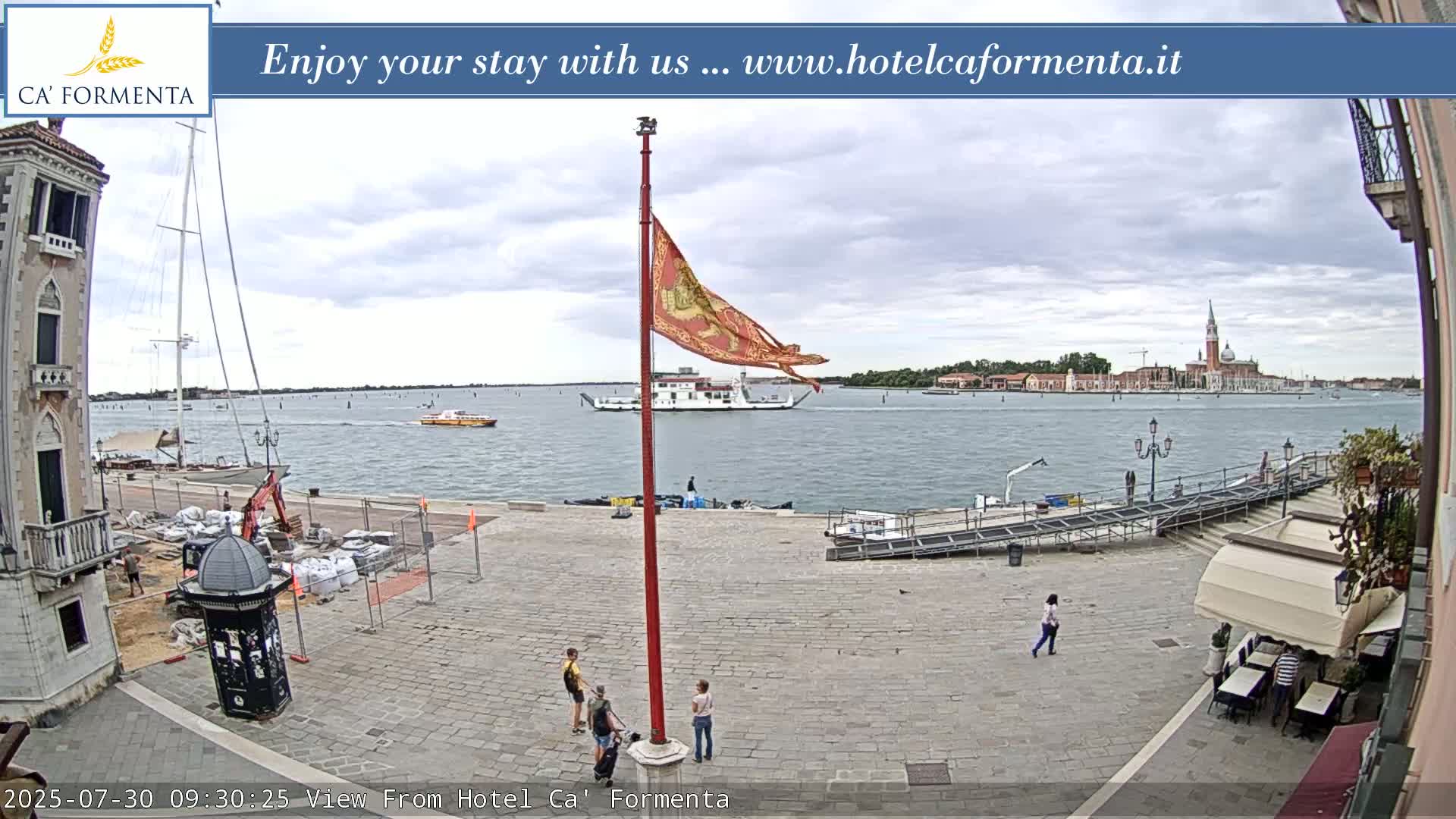 A partly cloudy day shows a Venetian plaza with a flagpole, people, and a view of a waterway with boats and a distant island.
