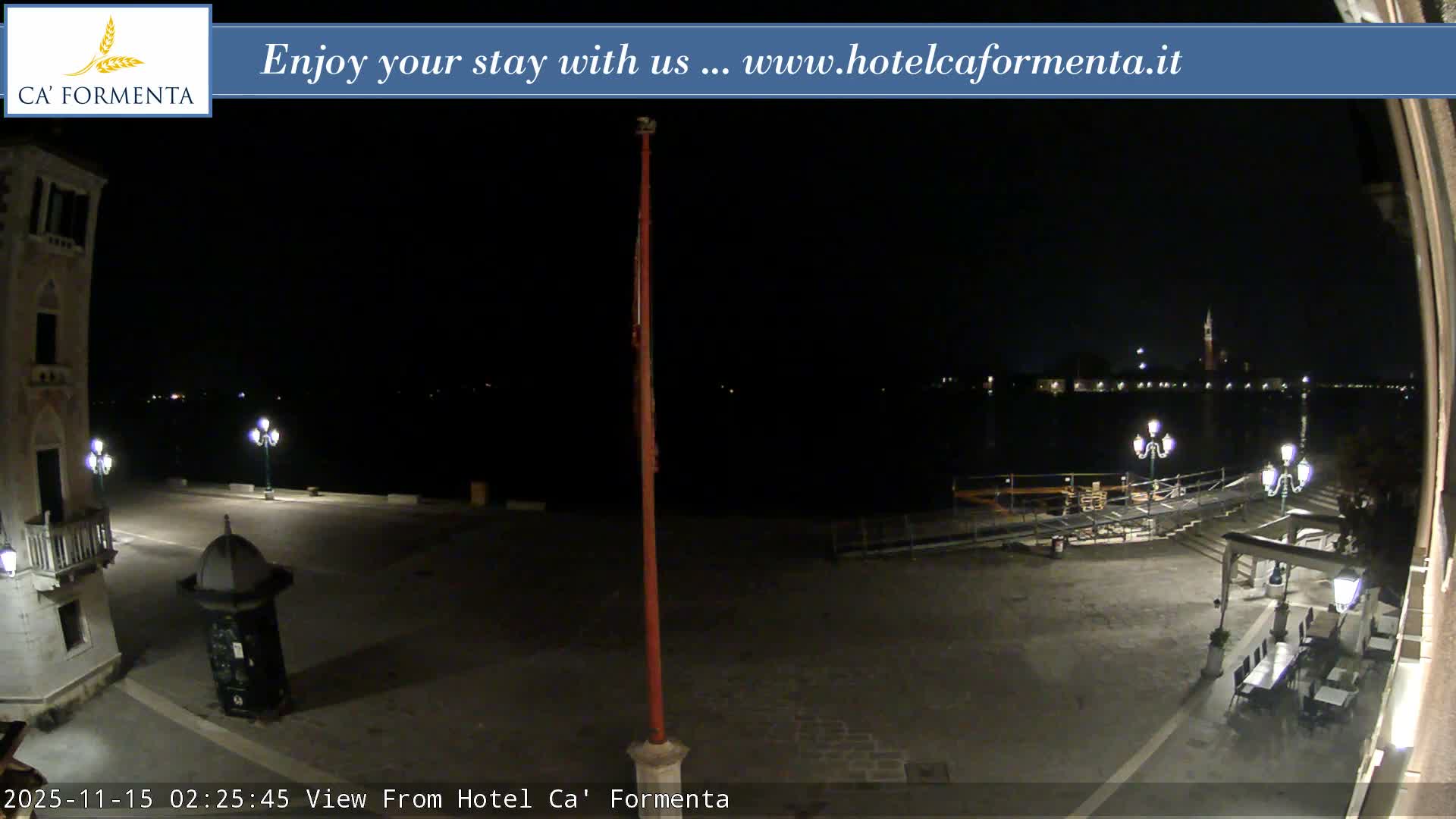 A clear night view captures a well-lit waterfront promenade with outdoor seating and streetlights, looking across dark water towards a distant city skyline featuring a prominent bell tower.
