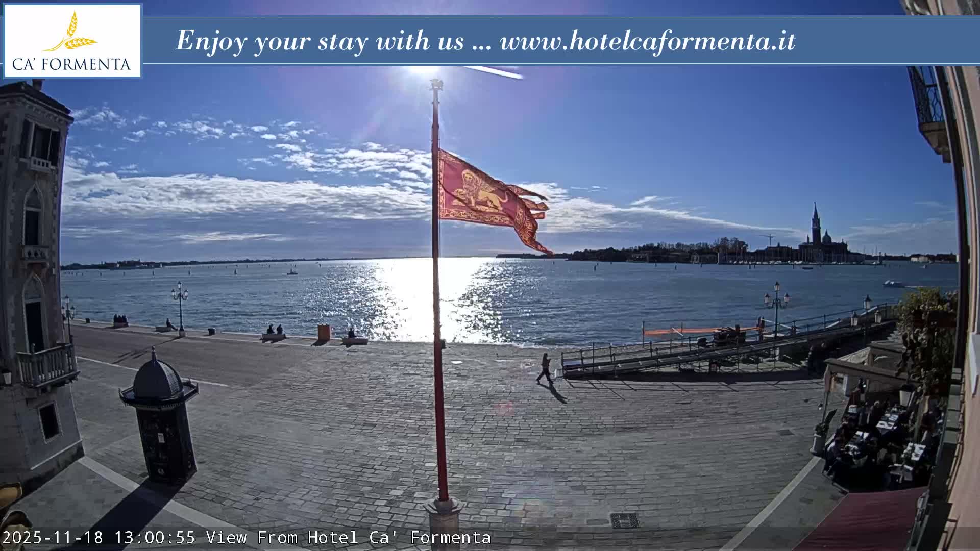 A clear night view captures a well-lit waterfront promenade with outdoor seating and streetlights, looking across dark water towards a distant city skyline featuring a prominent bell tower.
