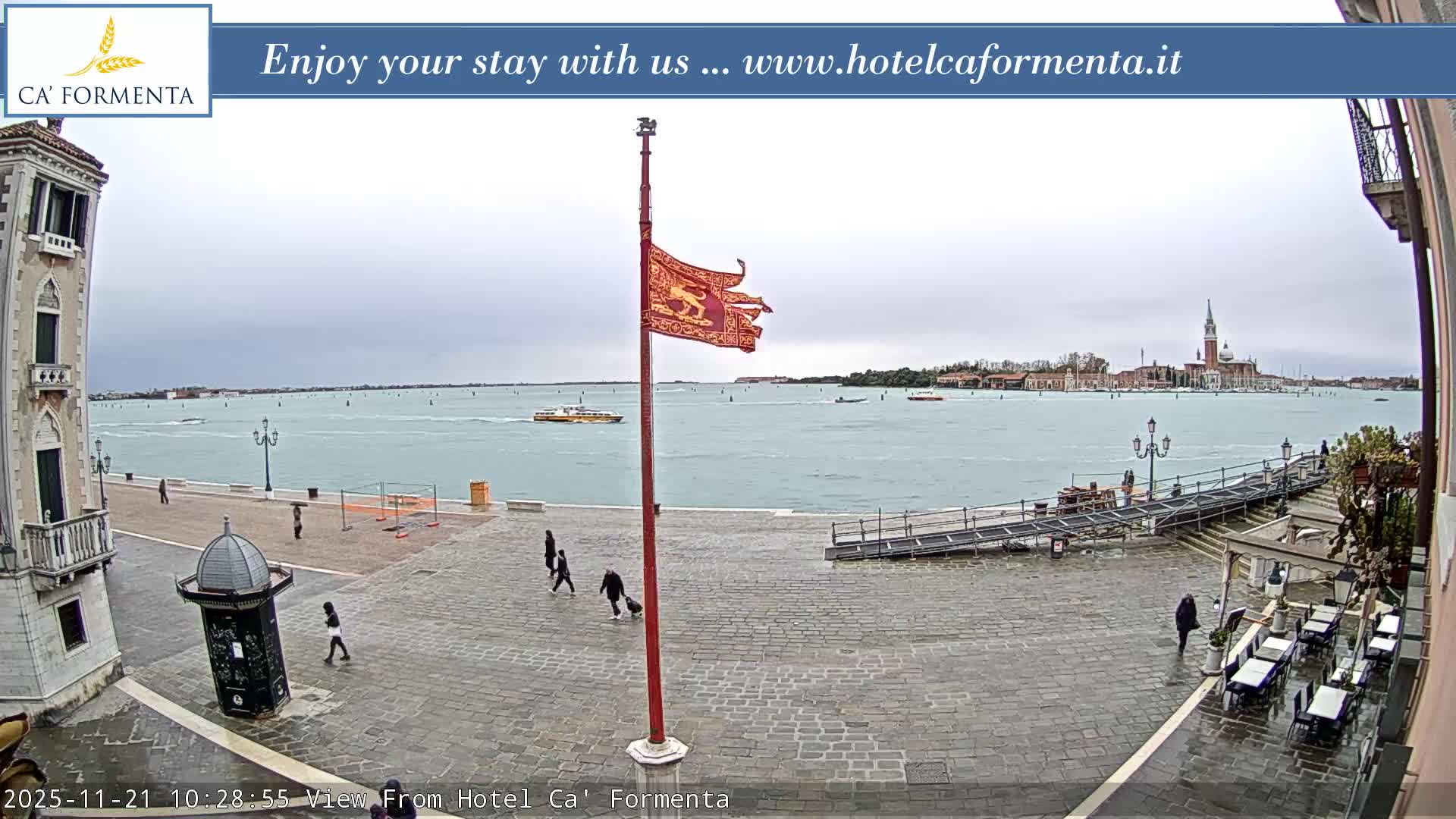 On a wet, overcast day in Venice, pedestrians walk across a cobblestone piazza featuring a flagpole flying a red and gold flag, overlooking the lagoon dotted with boats and the iconic San Giorgio Maggiore church in the distance.