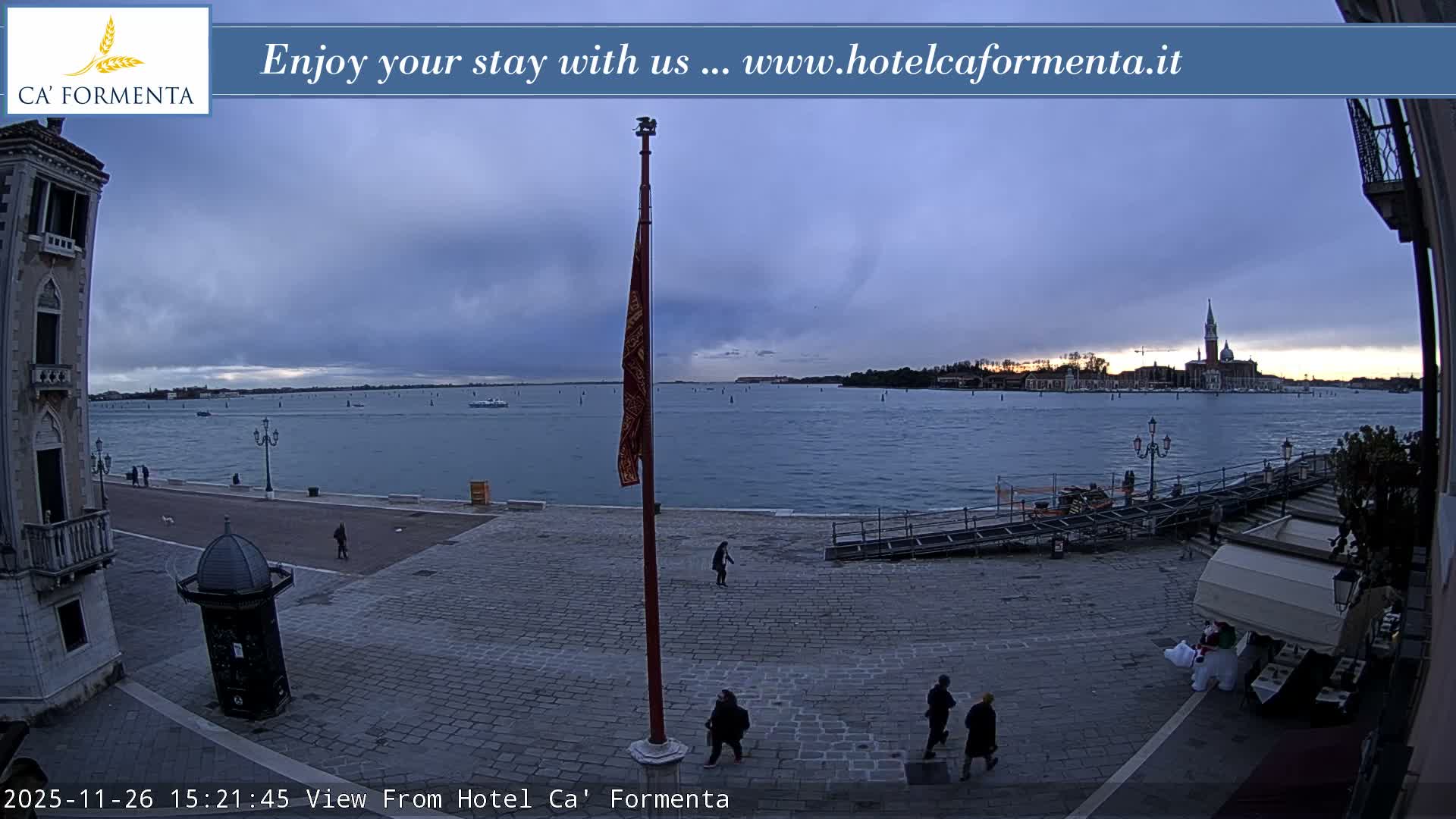 On a wet, overcast day in Venice, pedestrians walk across a cobblestone piazza featuring a flagpole flying a red and gold flag, overlooking the lagoon dotted with boats and the iconic San Giorgio Maggiore church in the distance.