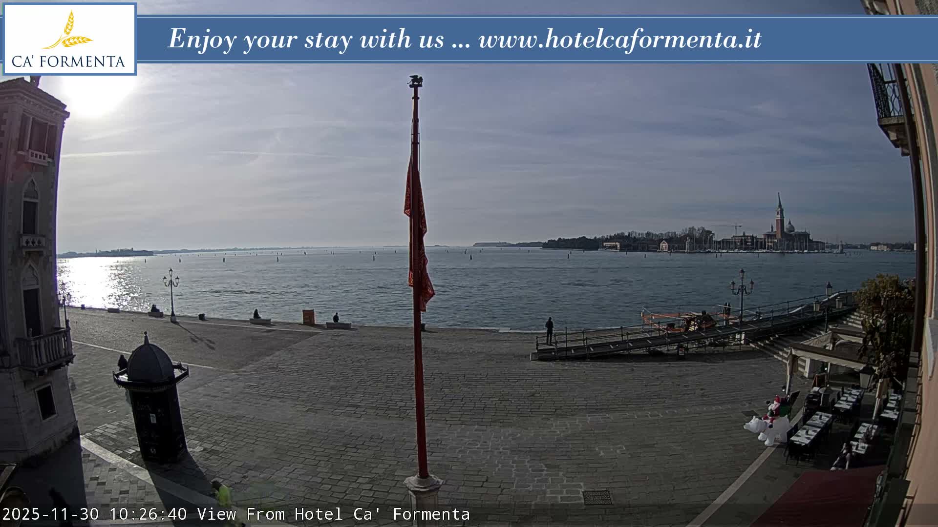 The image captures a sunny day with scattered clouds over a Venetian waterfront, showing a wide expanse of water with distant islands and the prominent San Giorgio Maggiore church, alongside a paved promenade with pedestrians and outdoor cafe seating.
