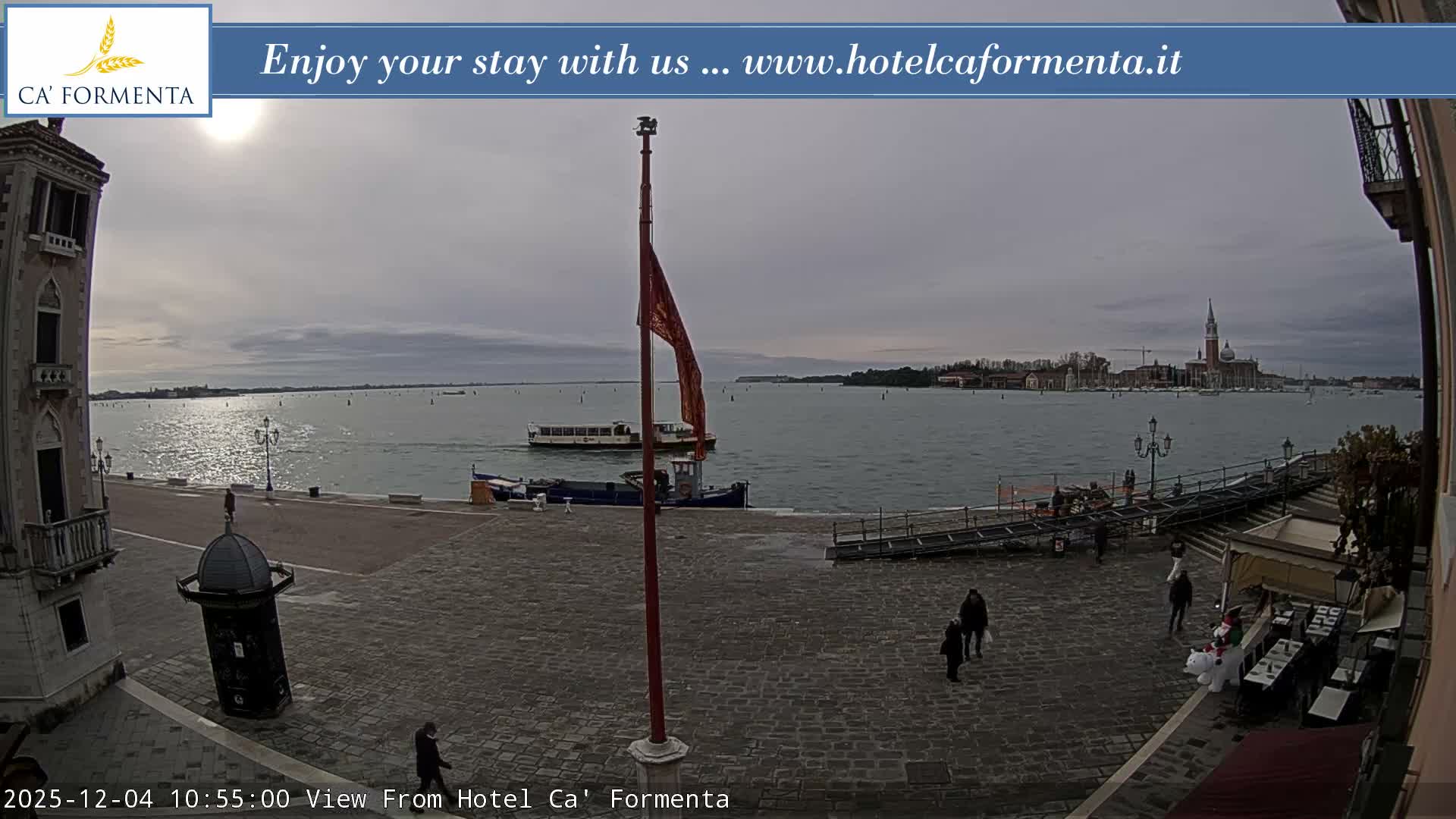 Under an overcast sky, the Venetian waterfront scene features a large red flag on a pole, numerous boats traversing the rippling water, a distant city skyline with a prominent church and bell tower, and people walking a cobblestone promenade past an outdoor cafe with a snowman figure.