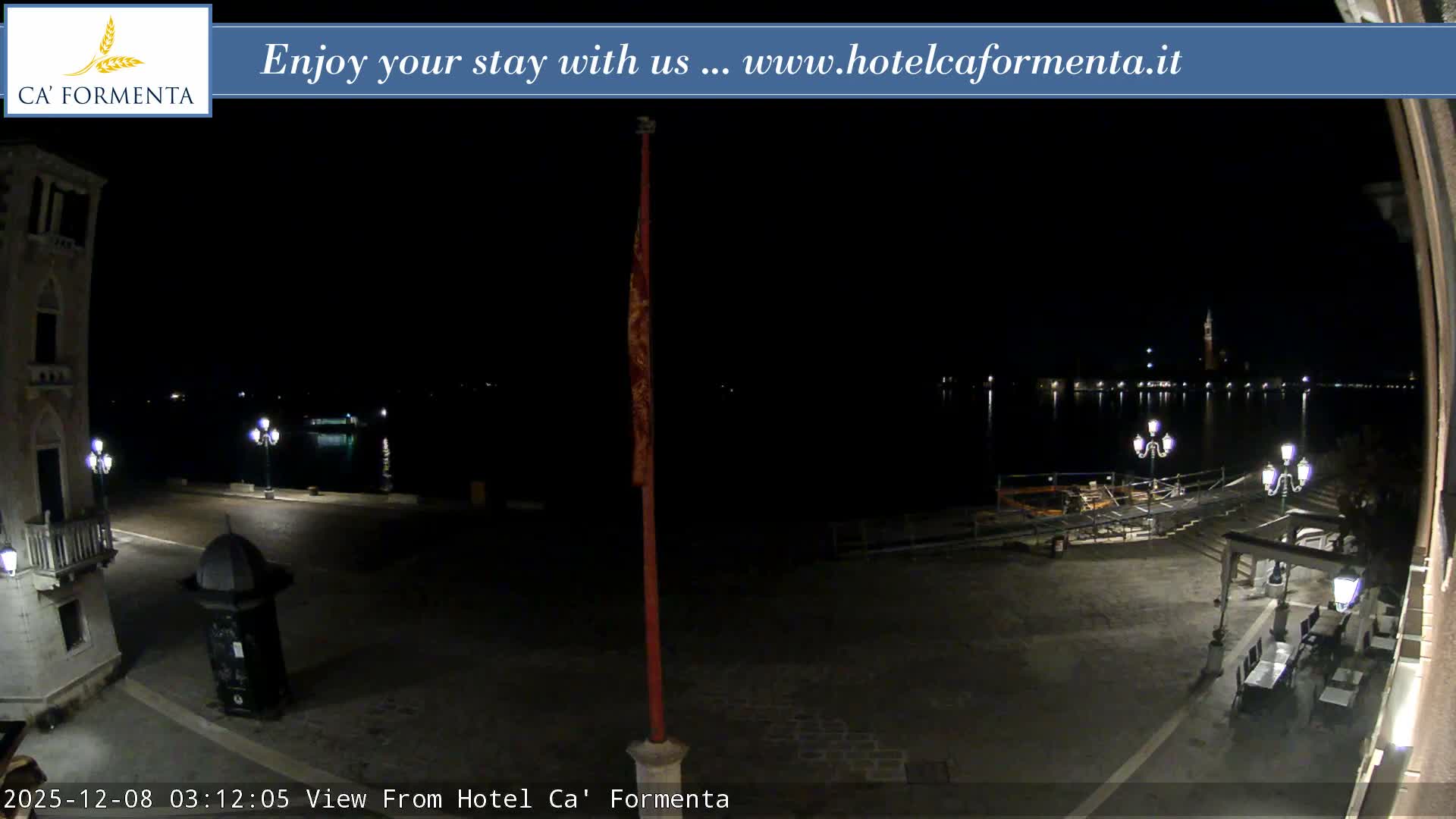 A clear night reveals a dimly lit waterfront plaza featuring ornate streetlights, outdoor seating under an awning, and a historic building, all overlooking calm water with city lights and a distant bell tower reflecting in the distance.