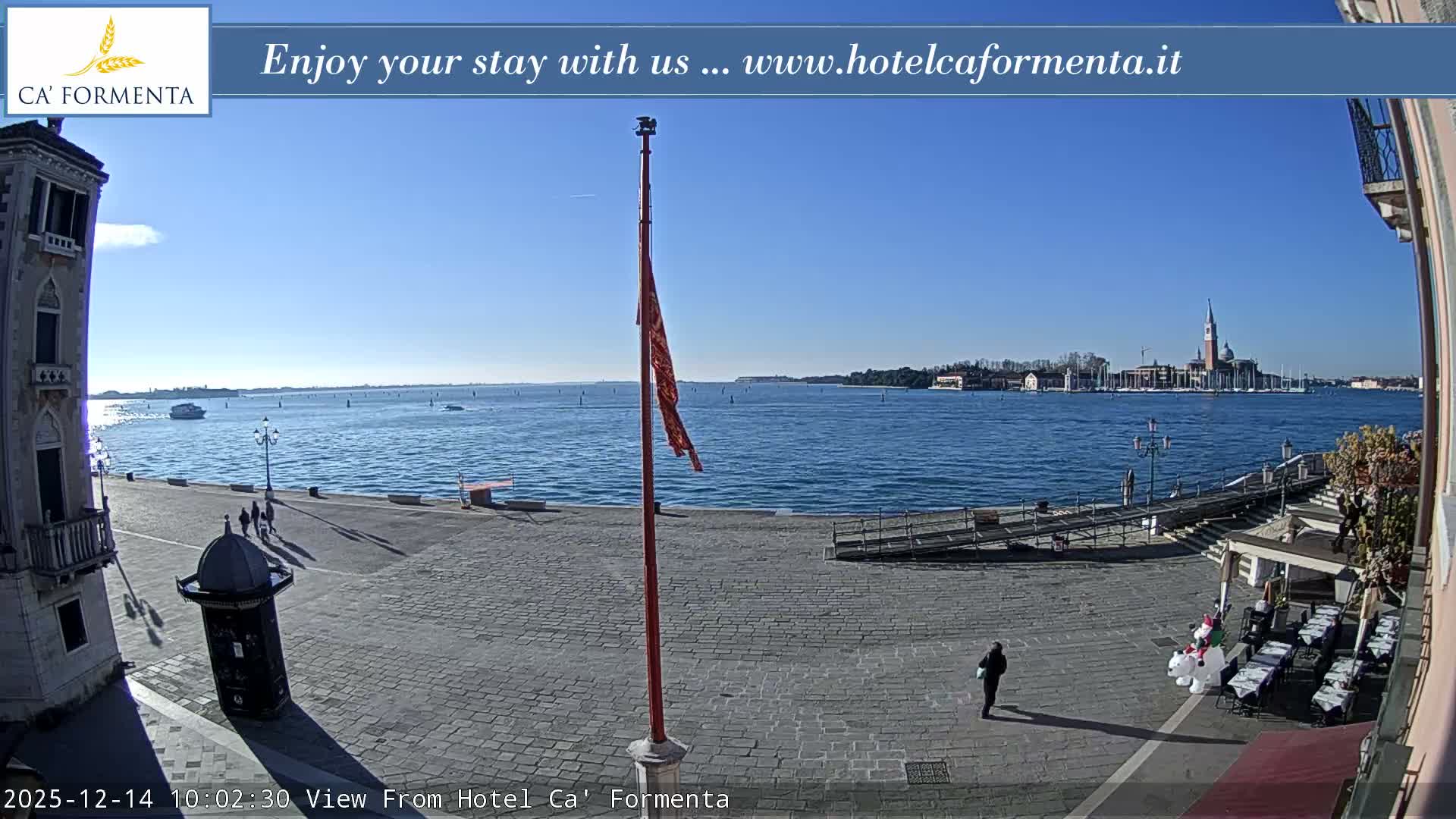 A clear night reveals a dimly lit waterfront plaza featuring ornate streetlights, outdoor seating under an awning, and a historic building, all overlooking calm water with city lights and a distant bell tower reflecting in the distance.