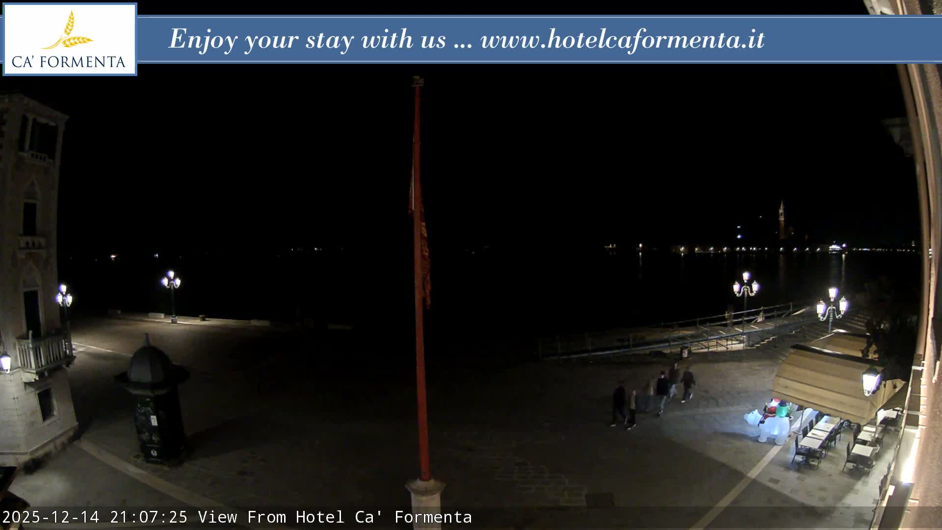 A clear night reveals a dimly lit waterfront plaza featuring ornate streetlights, outdoor seating under an awning, and a historic building, all overlooking calm water with city lights and a distant bell tower reflecting in the distance.