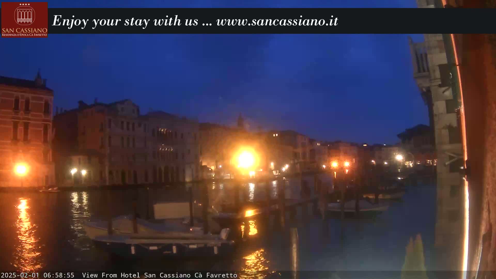 A nighttime view of a canal in Venice, with several buildings illuminated along its banks and boats moored on the water under a dark blue sky.