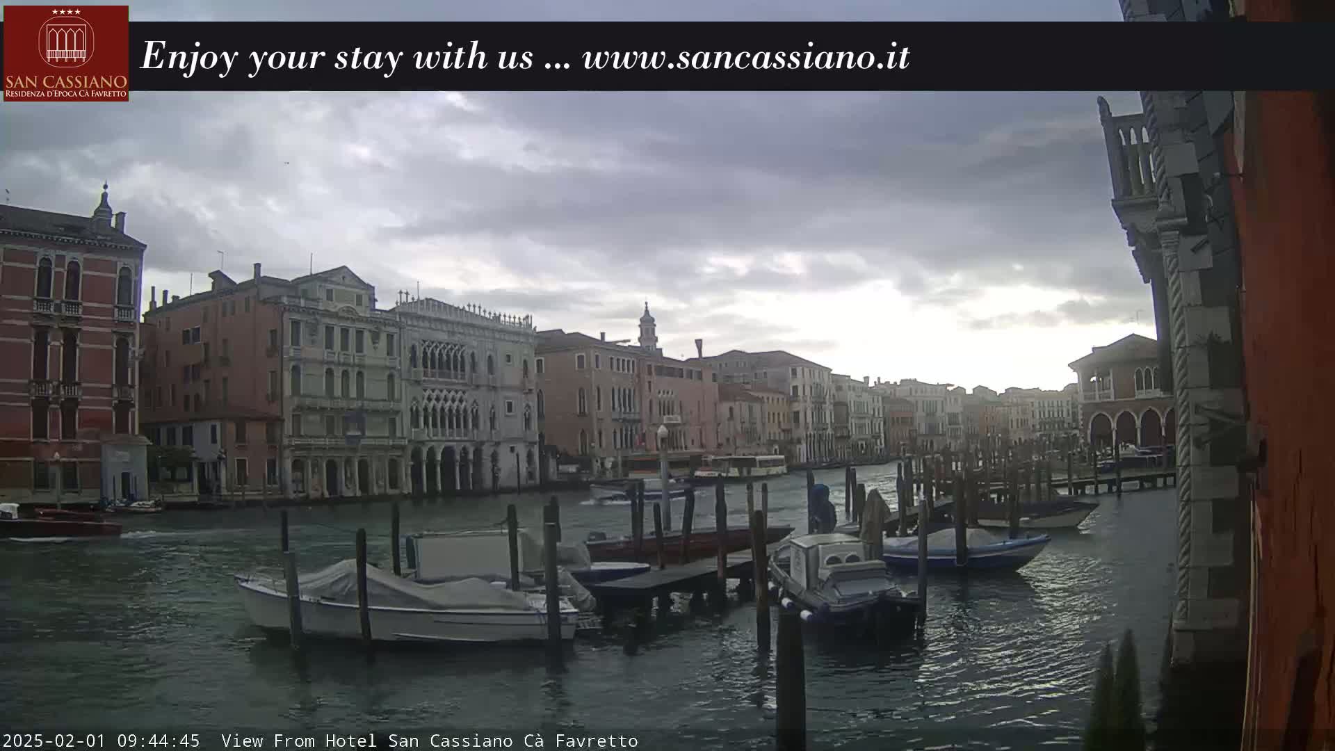 A canal in Venice, under an overcast sky, shows several boats moored along a dock in front of historic buildings.
