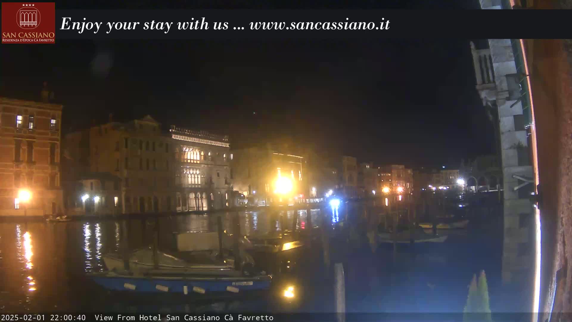 A nighttime view of a canal in Venice, Italy, showing illuminated buildings and boats along the waterway under a clear, dark sky.